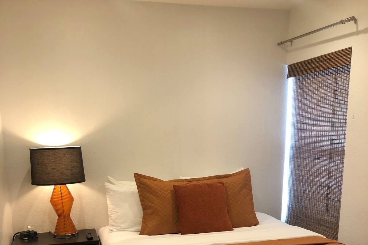 A neatly made bed with a brown bedspread and pillows, a bedside table with a lamp, and a window with a mesh screen.