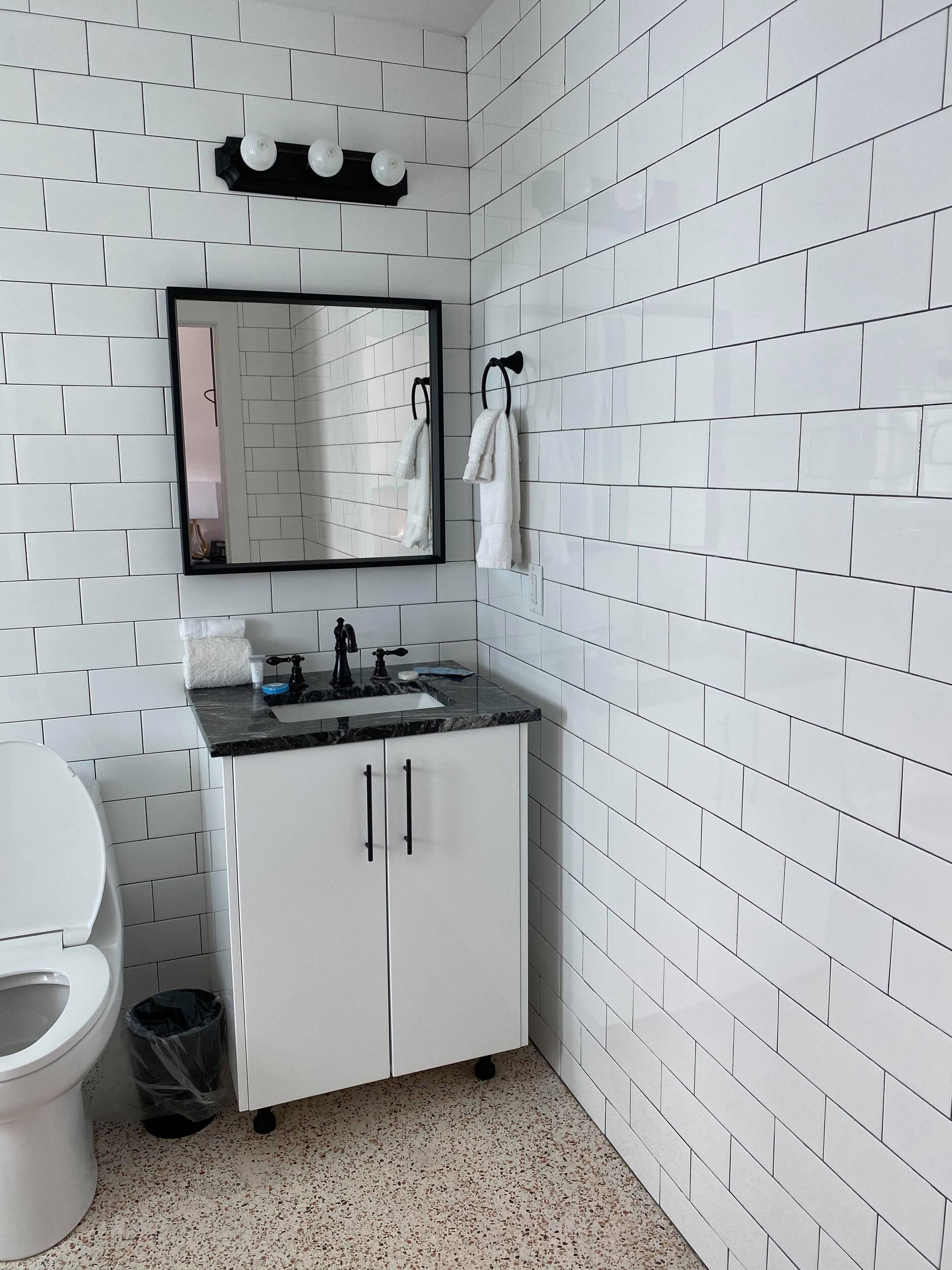 Ein Badezimmer mit weißen Fliesen, einem weißen Waschbecken mit schwarzer Arbeitsplatte, einem Spiegel über dem Waschbecken und einer Toilette links.