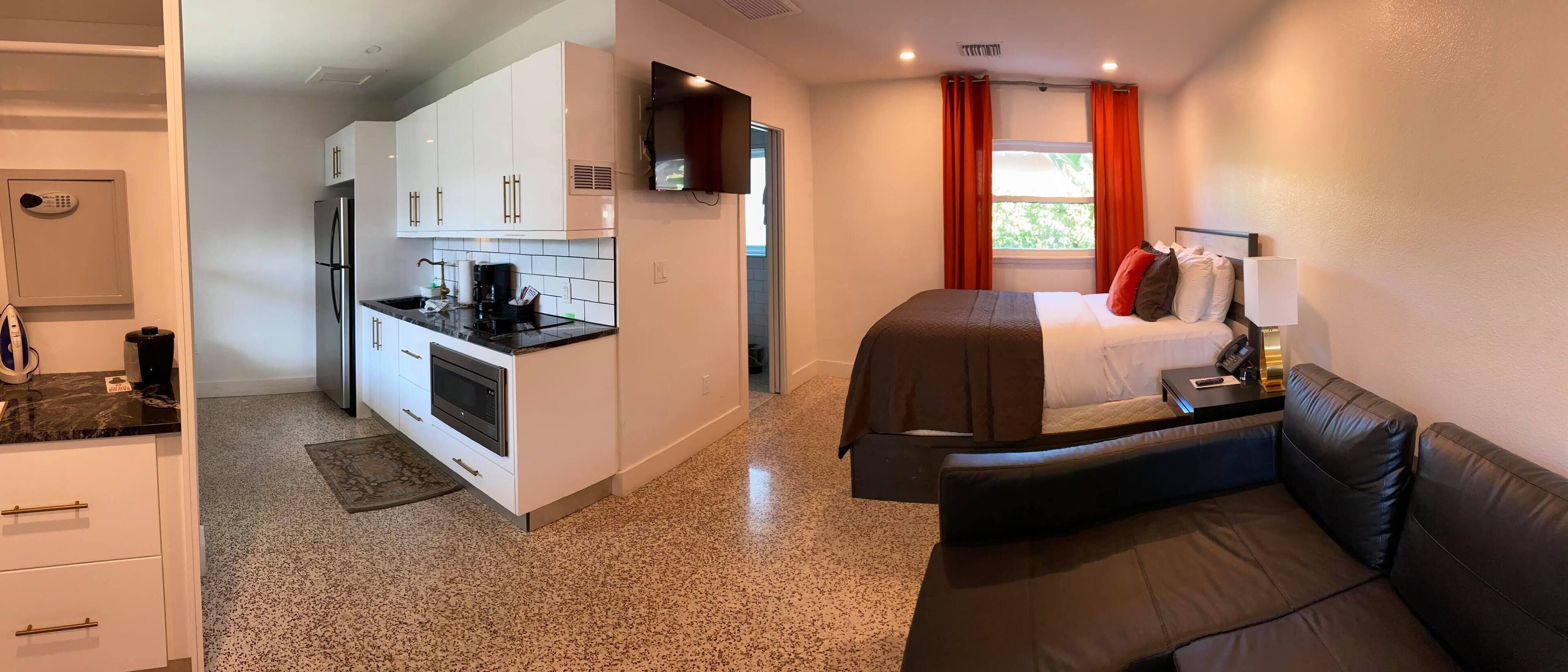 A compact hotel room with a kitchenette, a bed with a headboard, a bedside table with a lamp, and a view of the outdoors through a window.