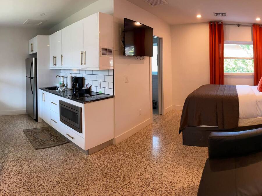 A compact hotel room with a kitchenette, a bed with a headboard, a bedside table with a lamp, and a view of the outdoors through a window.
