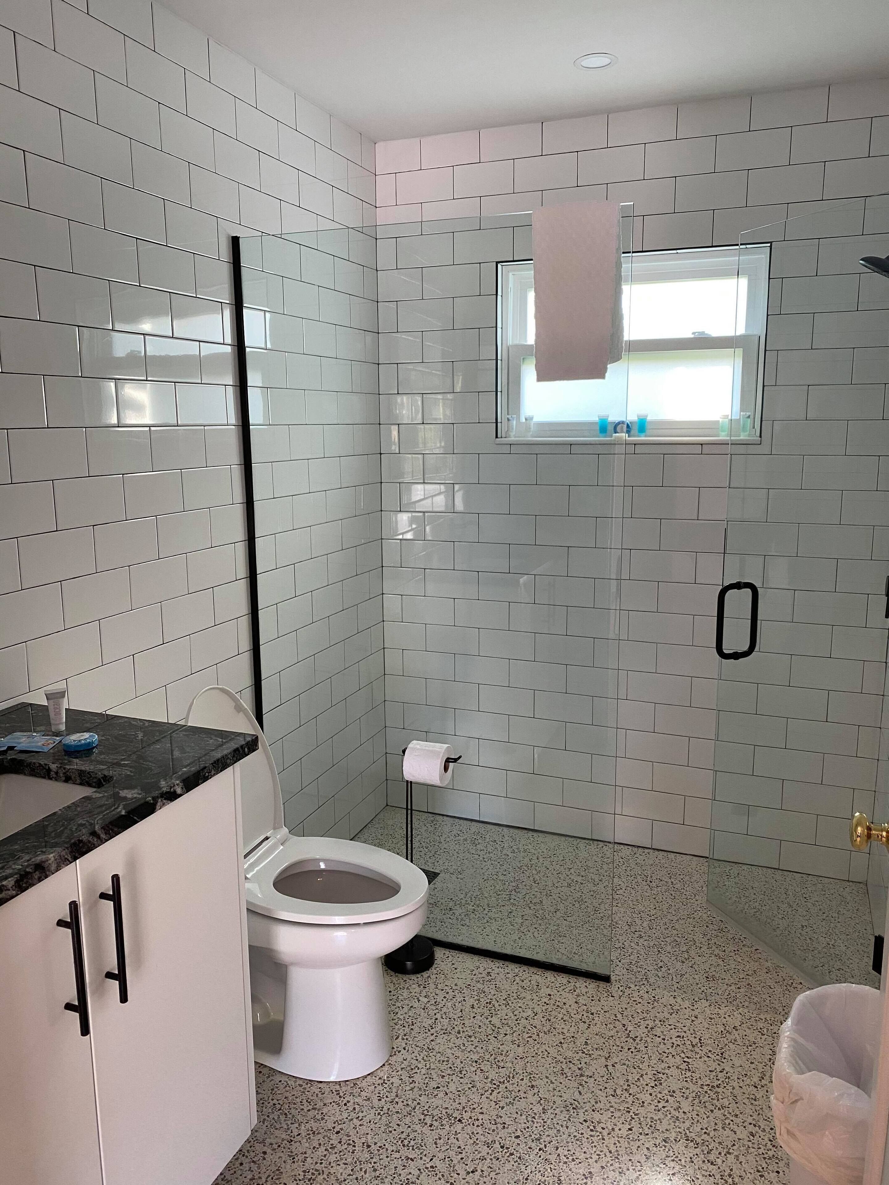 A bathroom with white tiled walls, a white toilet, a sink with a black countertop, and a window with a towel hanging.