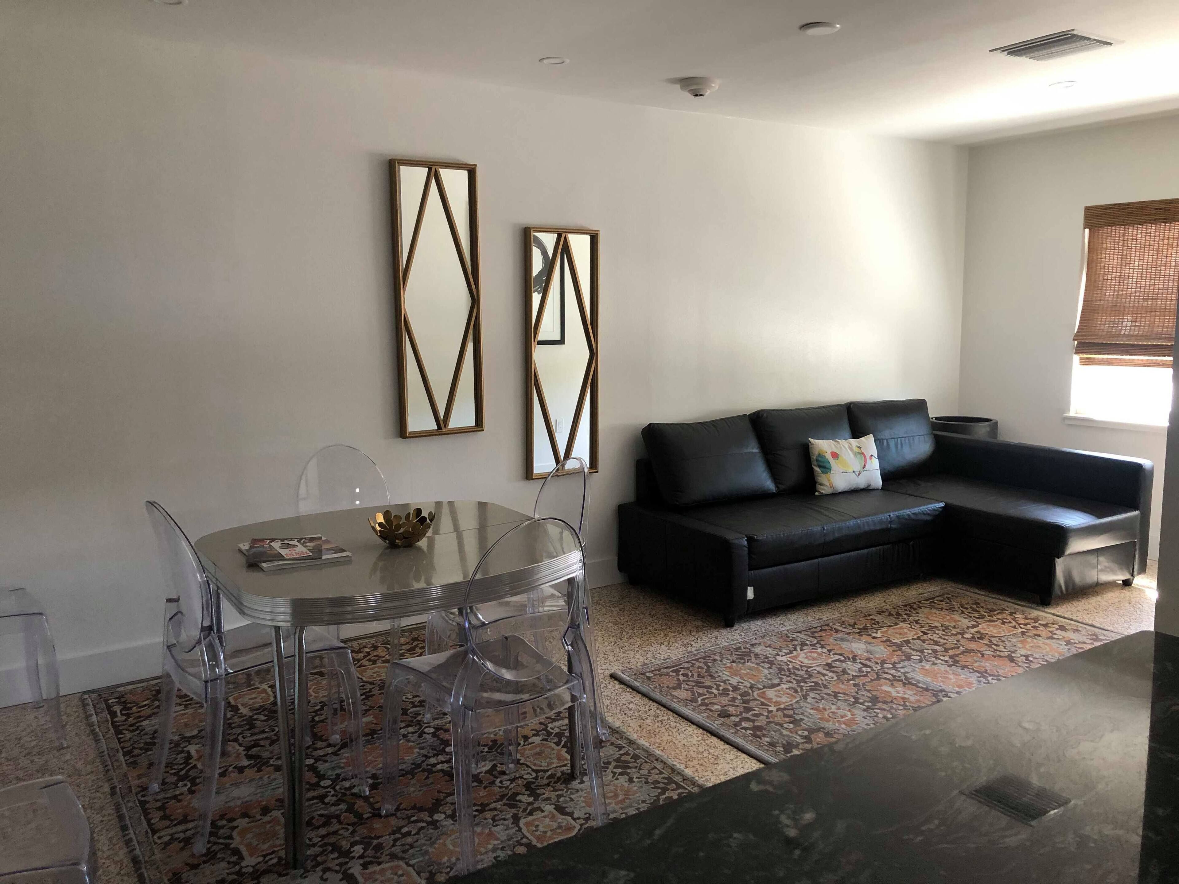 A modern living room with a glass dining table, clear chairs, a black leather sofa, and two geometric wall mirrors.