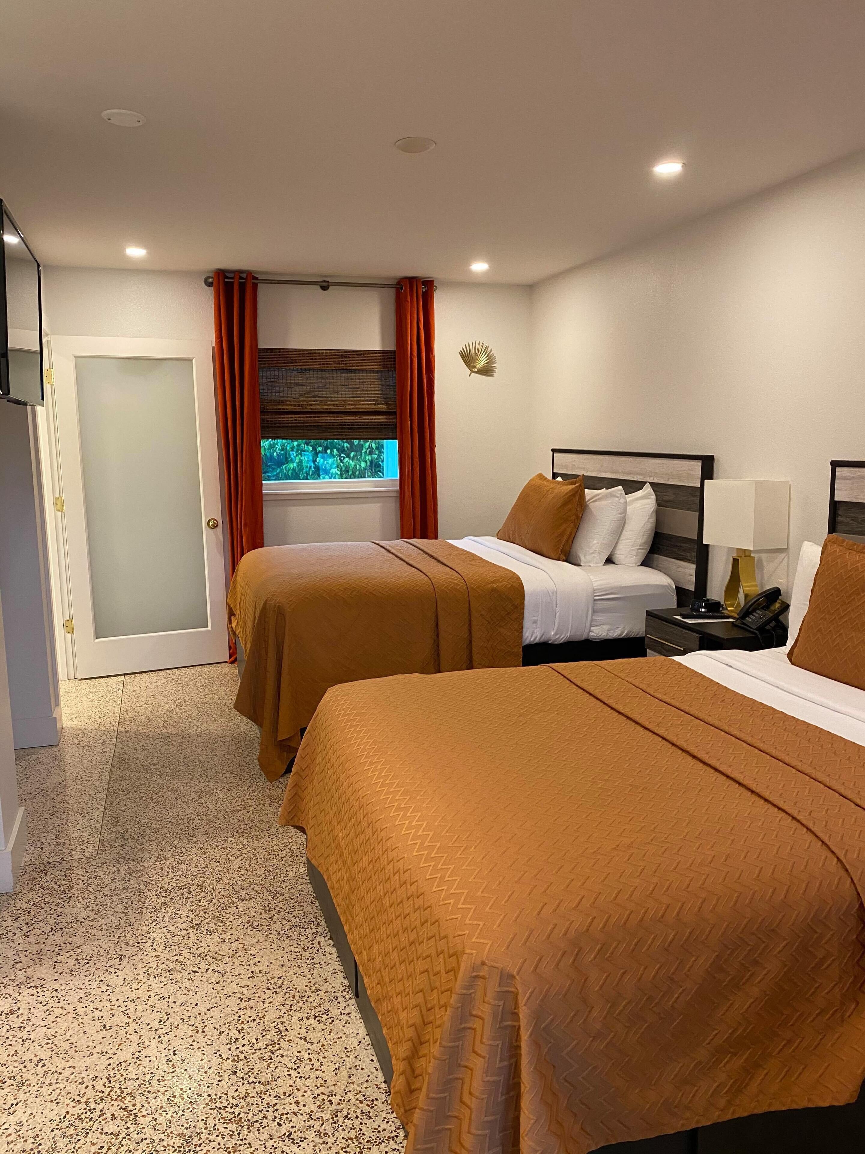 Habitación de hotel con dos camas, una ventana con cortinas, una mesita de noche con lámpara y un ventilador de pared.