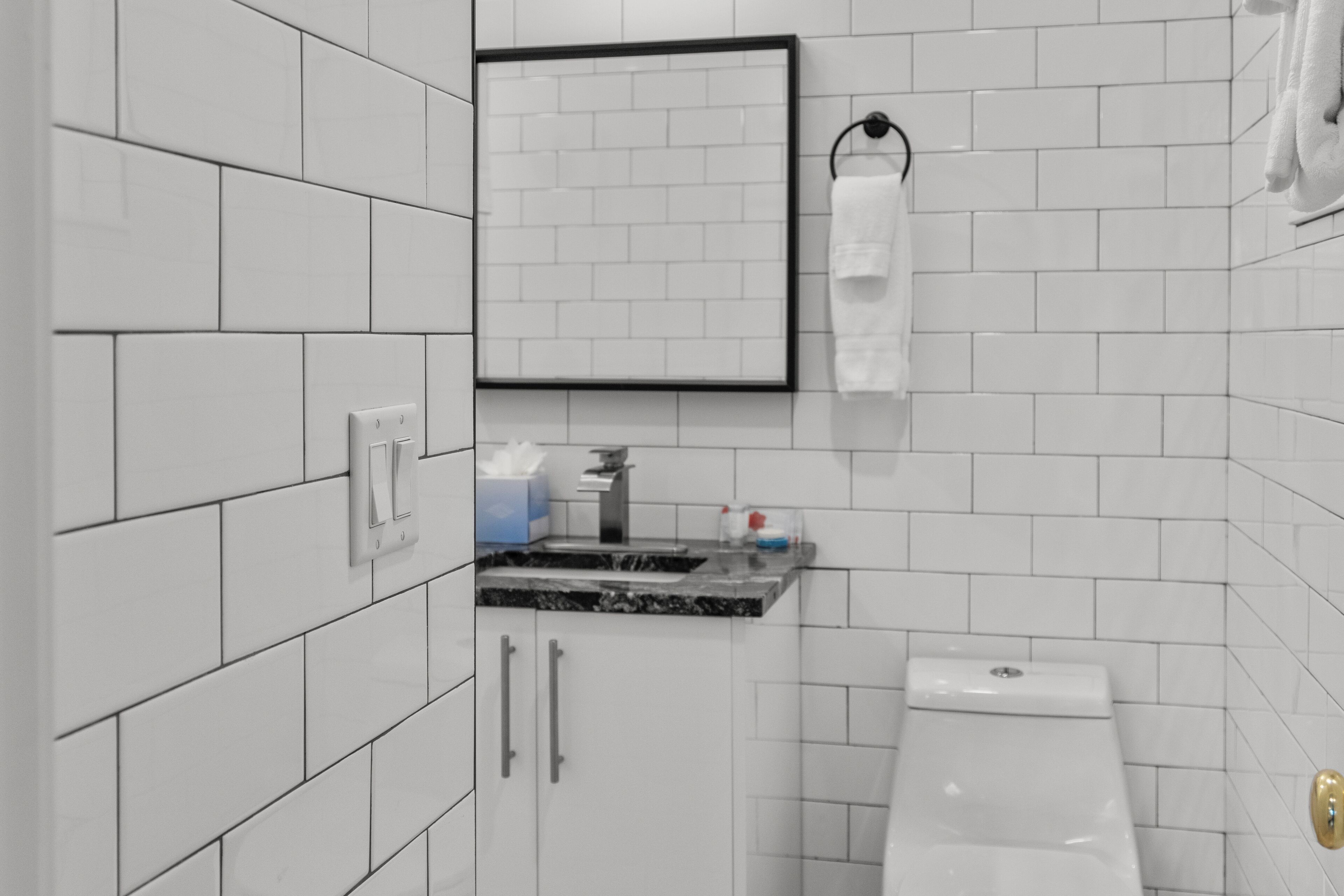 A bathroom with white tiled walls, a sink with a marble countertop, a toilet, and a framed mirror.