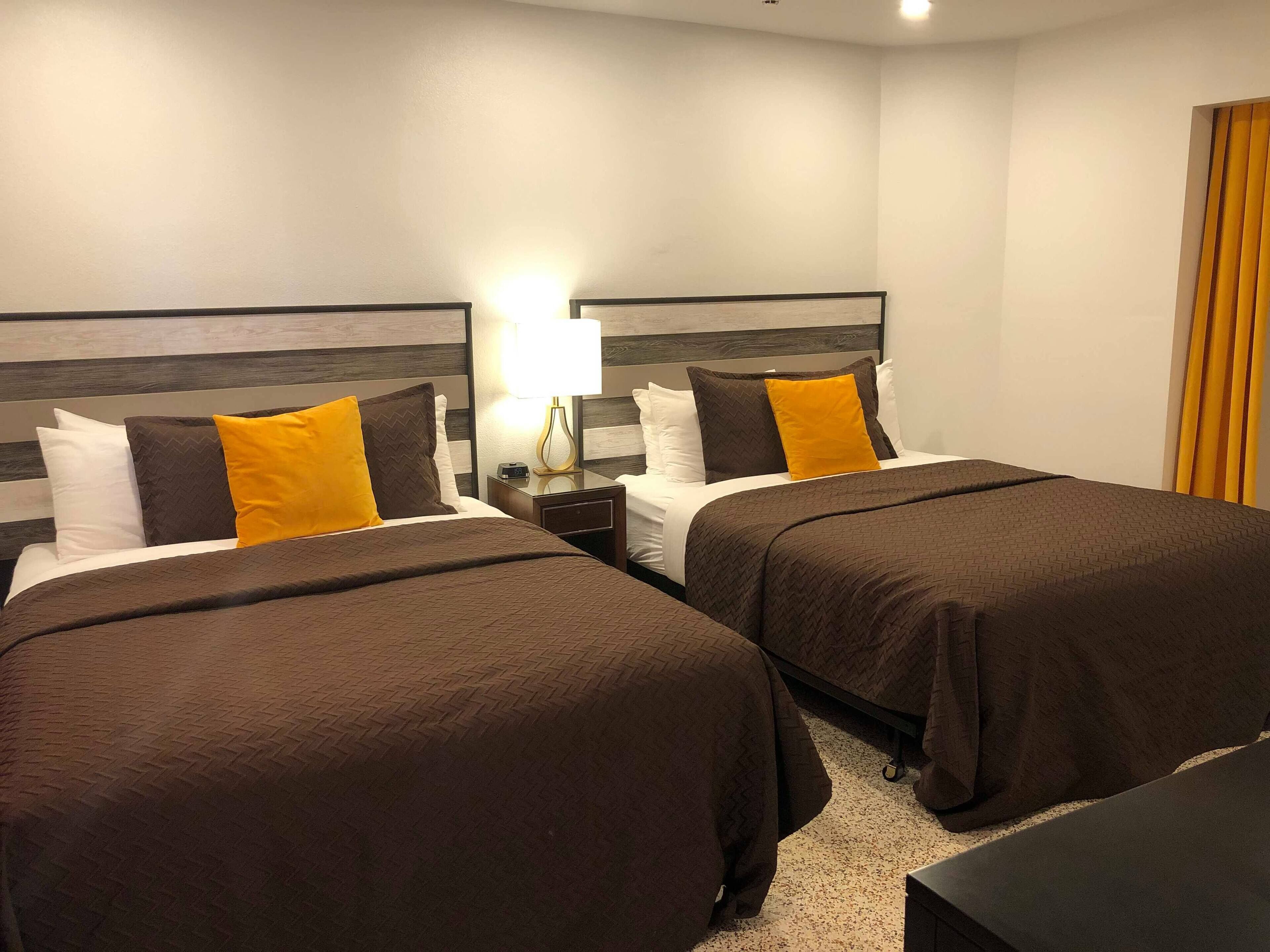 A hotel room with two beds, each with a brown bedspread and pillows, a bedside table with a lamp, and a wooden headboard.