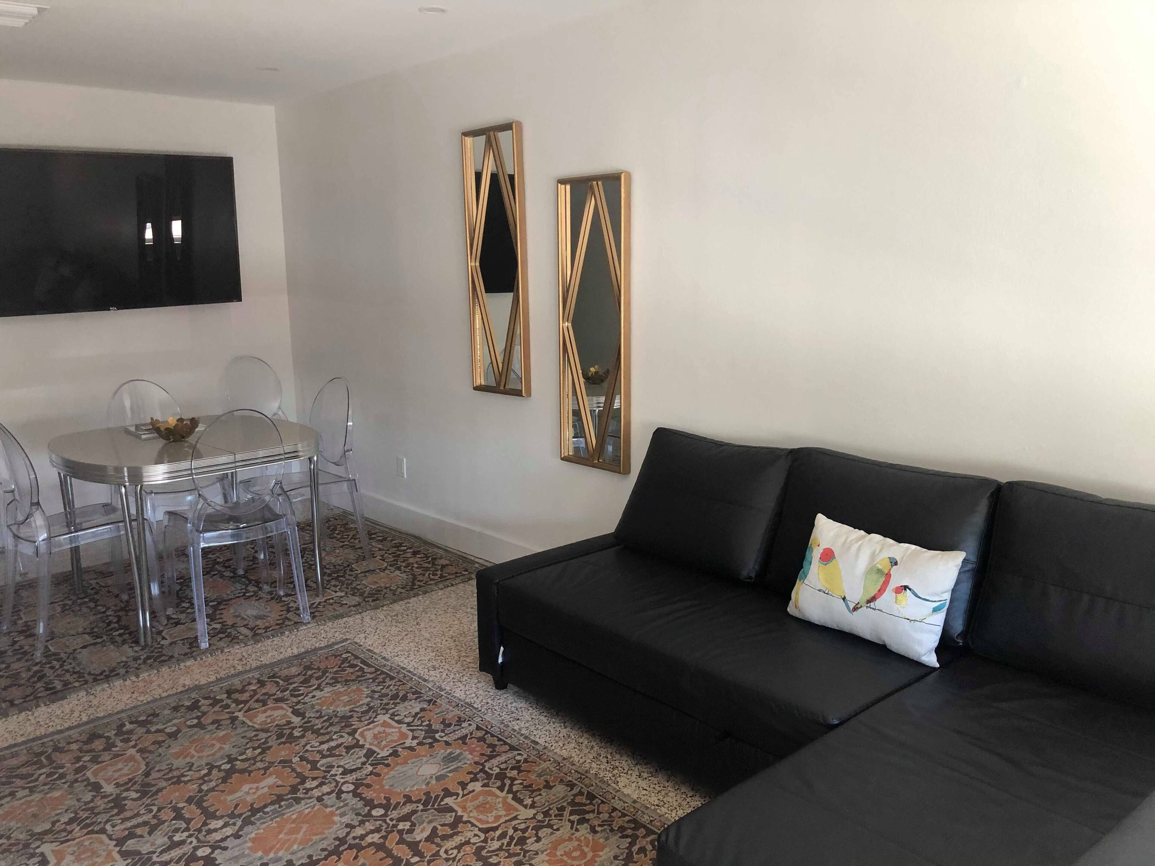 A living room with a black leather sofa, a glass dining table with chairs, a patterned rug, and two wall-mounted mirrors.