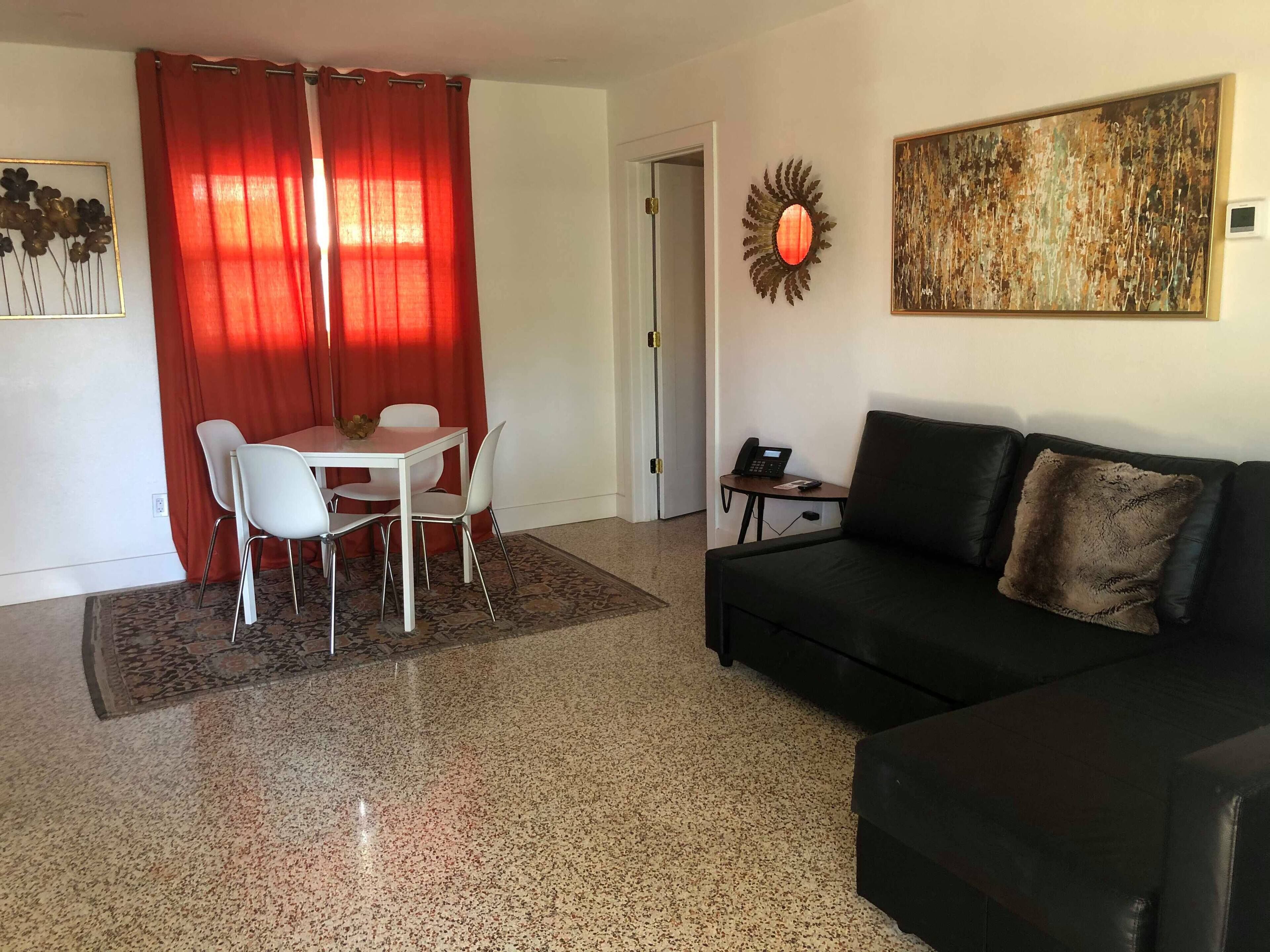 A living room with a black sofa, a dining table with white chairs, a red curtain, and a wall-mounted phone.