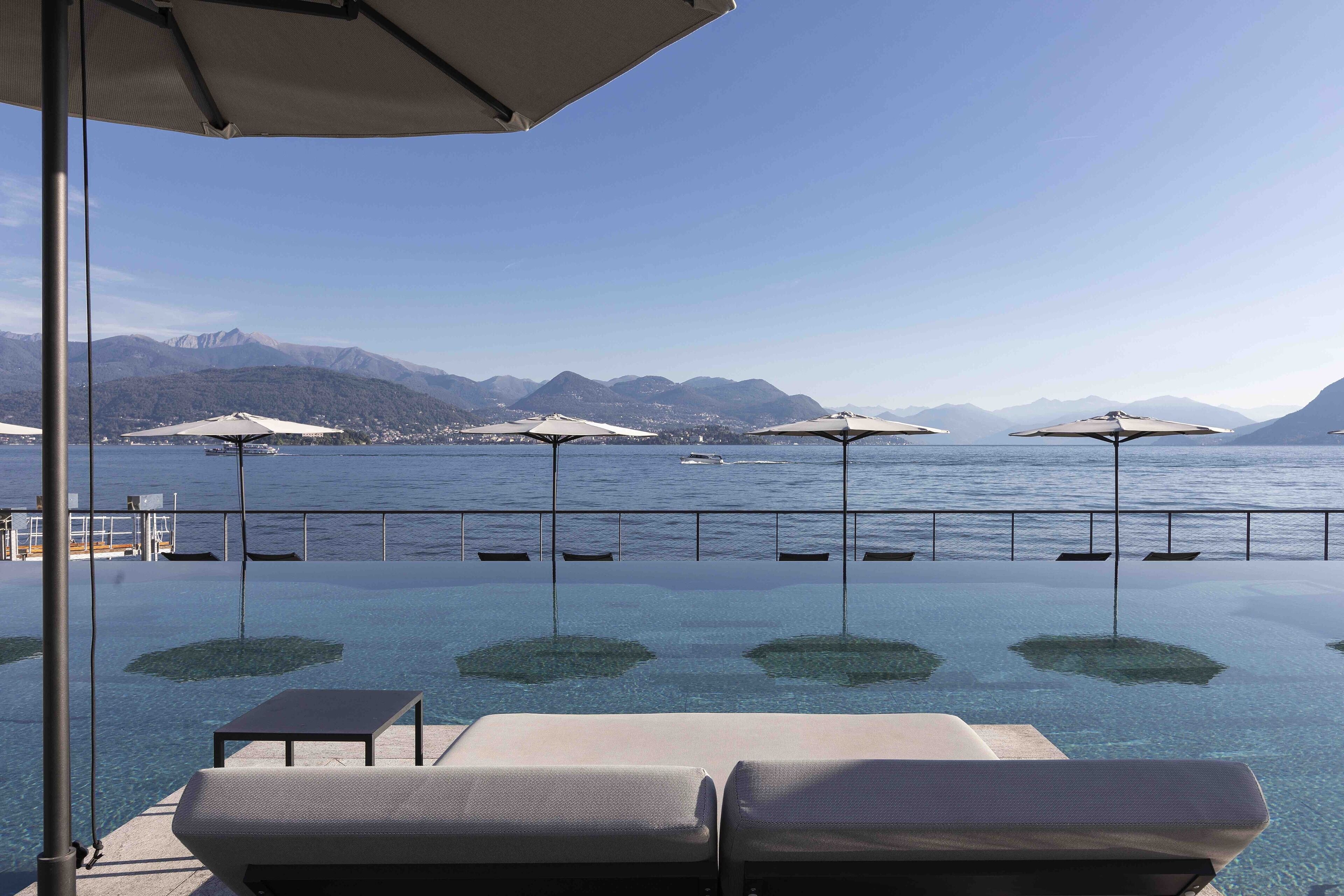Una piscina infinita con sillas de descanso y sombrillas, con vistas a un lago y montañas al fondo.