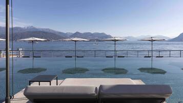 Una piscina infinita con sillas de descanso y sombrillas, con vistas a un lago y montañas al fondo.