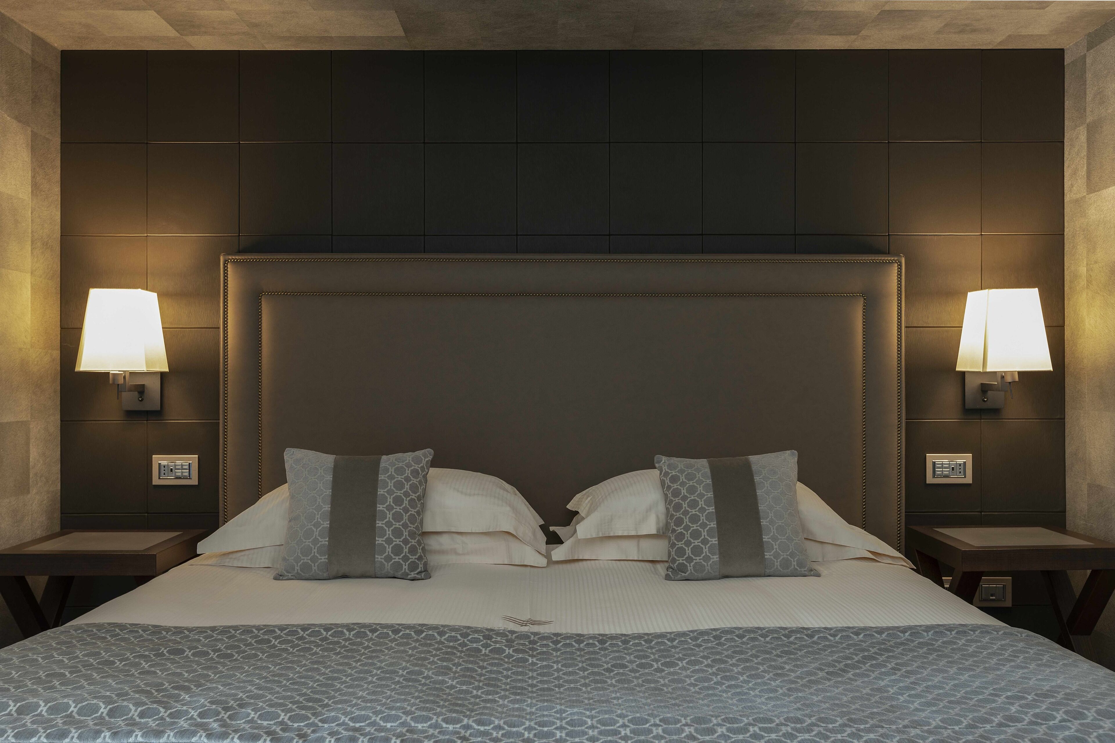 A neatly made bed with a patterned bedspread, two pillows, and a headboard. There are two bedside tables with lamps and a wall-mounted control panel.