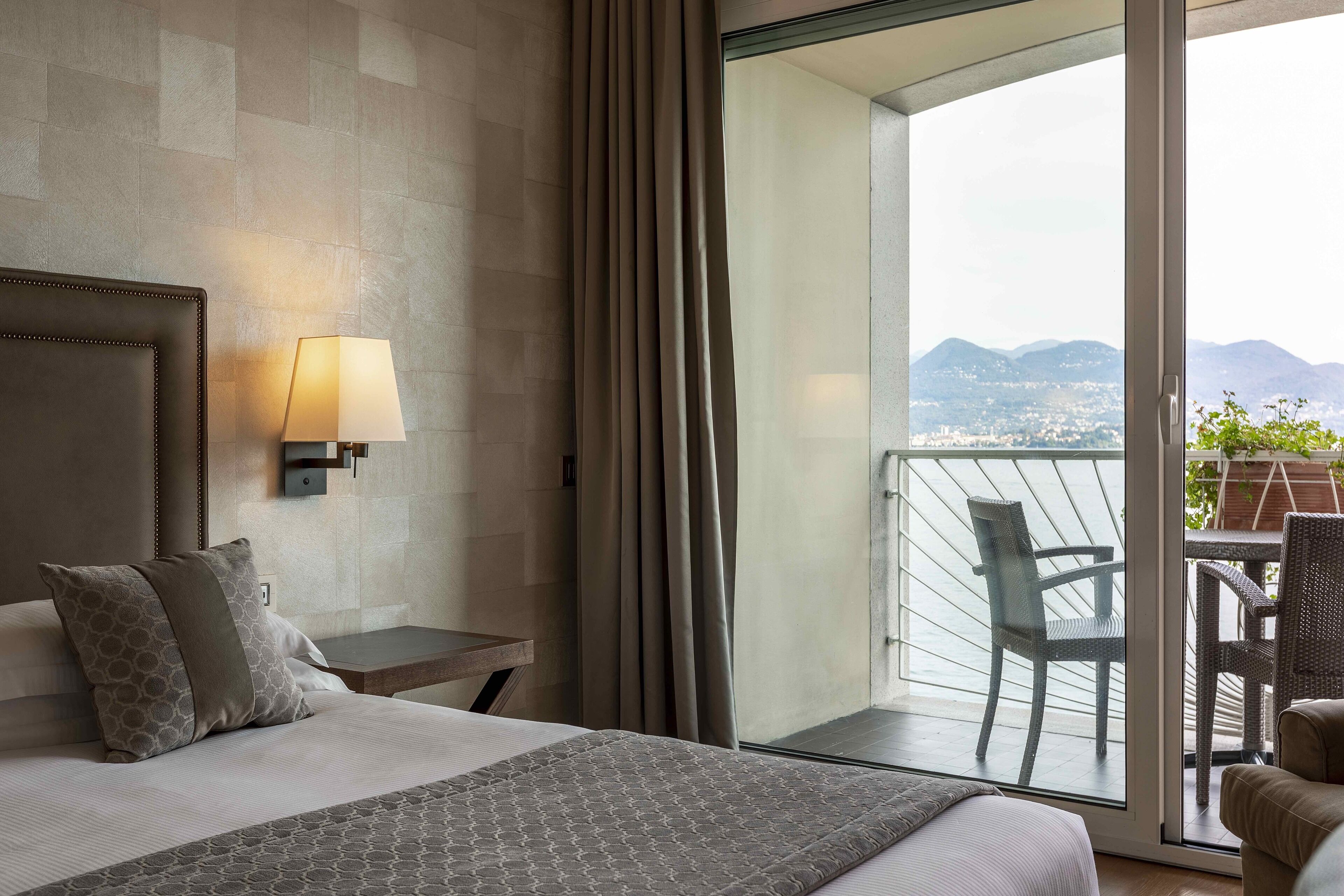 A hotel room with a large bed, a nightstand, a lamp, and a view of the city and mountains through a glass door.