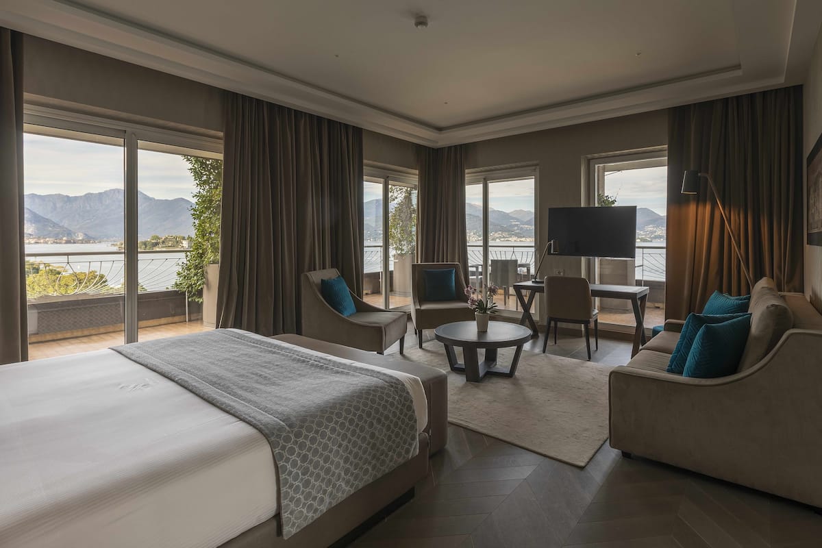 Ein Hotelzimmer mit einem großen Bett, zwei Sesseln, einem Couchtisch und einem Fernseher. Das Zimmer verfügt über große Fenster mit Blick auf Berge und einen See.