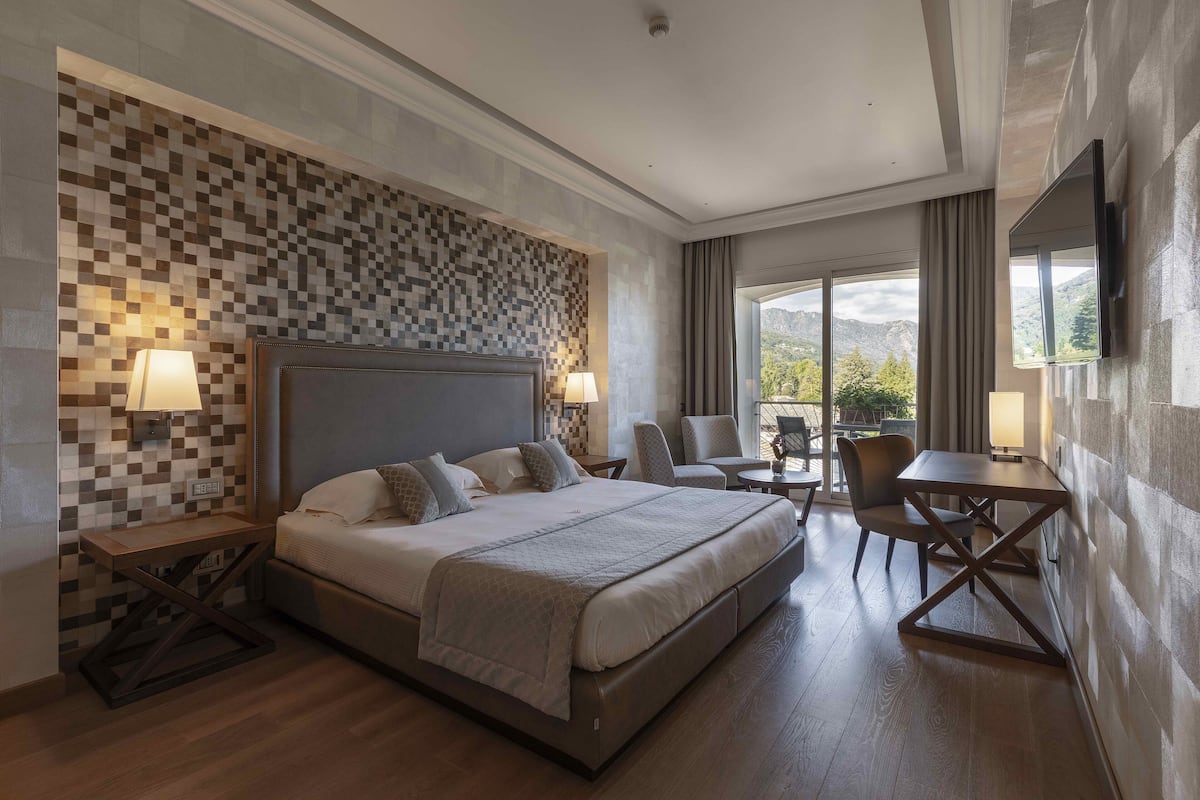 Ein modernes Hotelzimmer mit einem großen Bett, einem Schreibtisch und einem Sessel. Durch die Fenster ist eine Bergblick zu sehen.