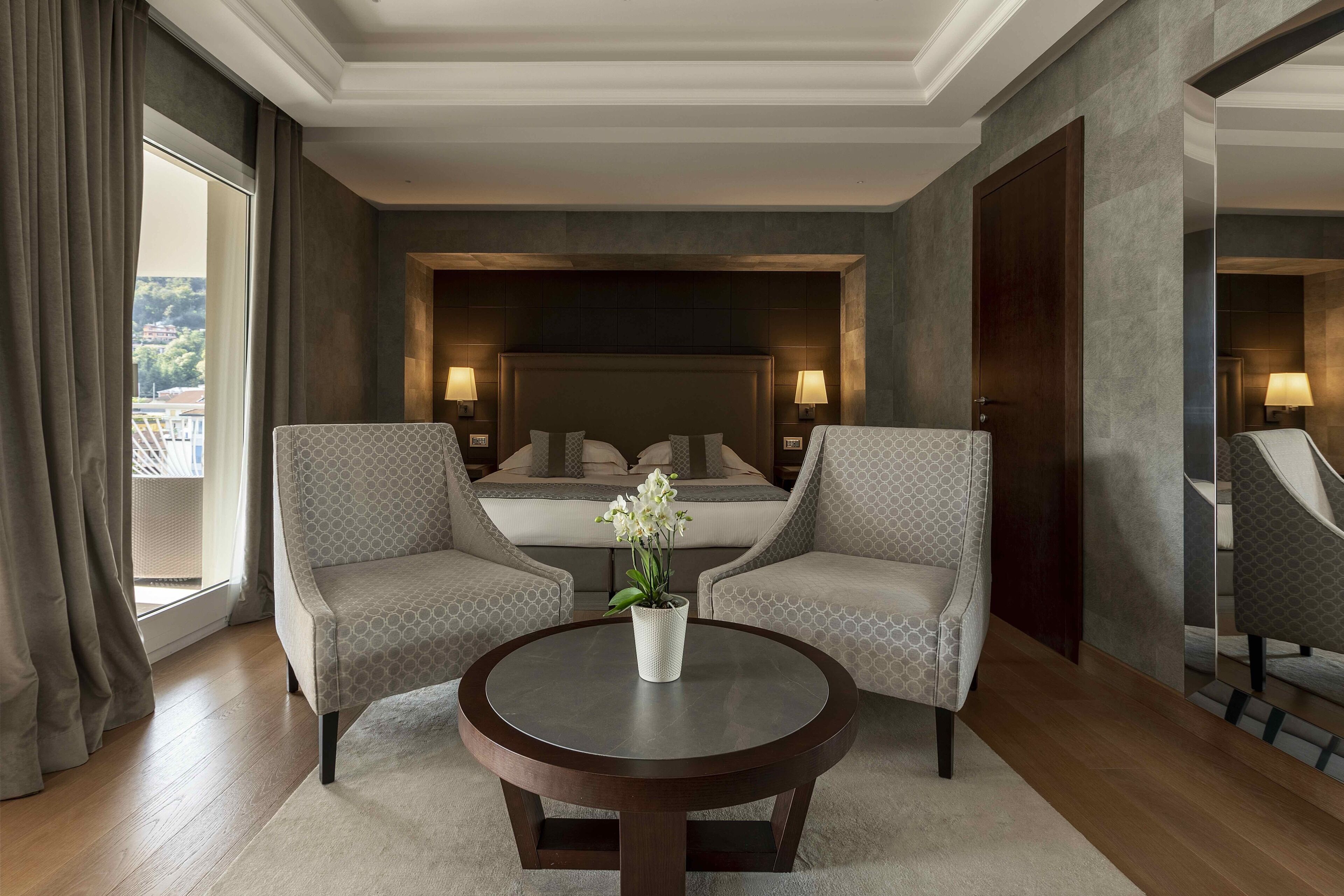 A hotel room with a bed, two armchairs, a round table with a vase, and a view of the outdoors.