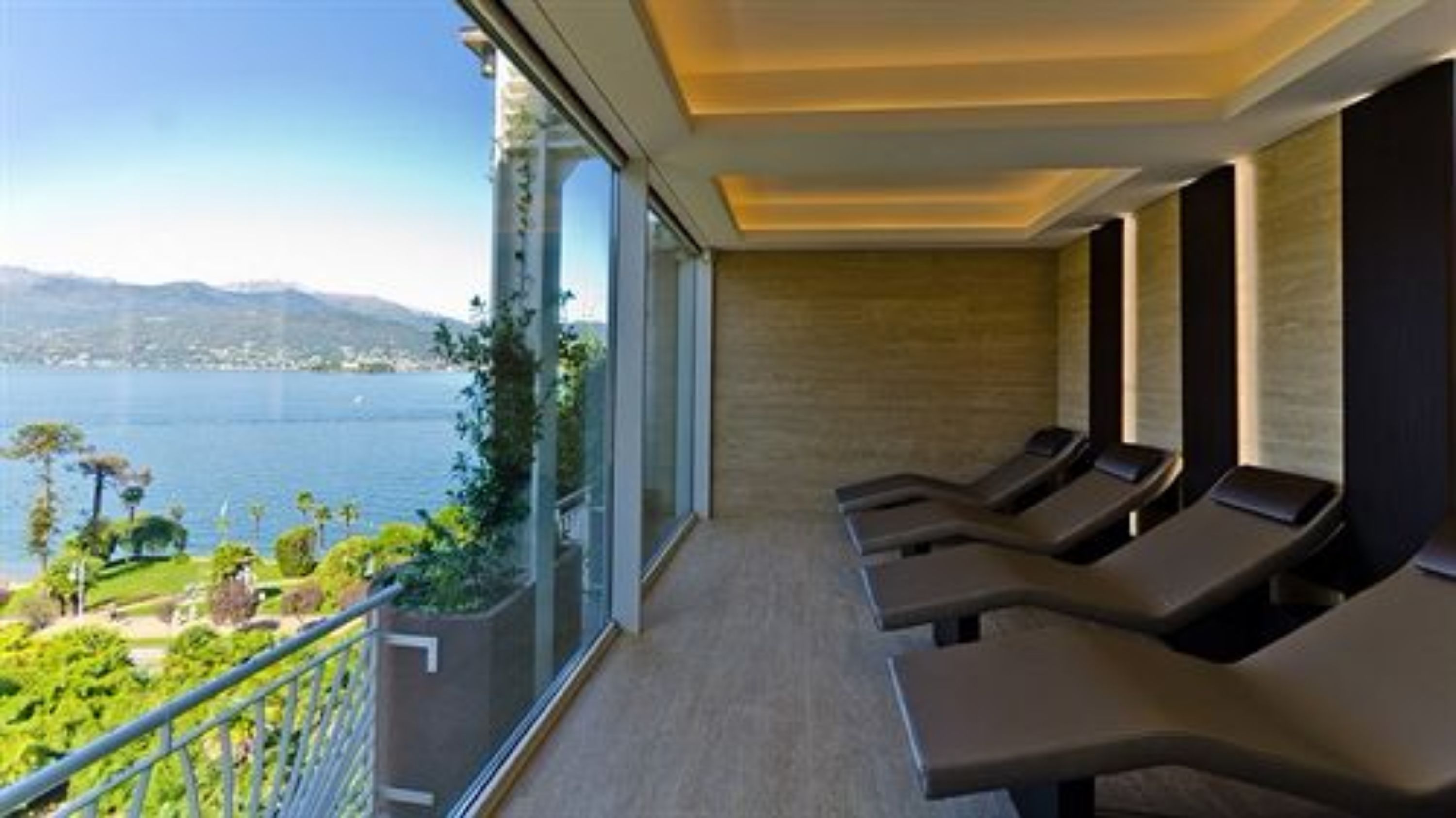 A balcony with a view of a lake and mountains, equipped with sun loungers and a glass railing.