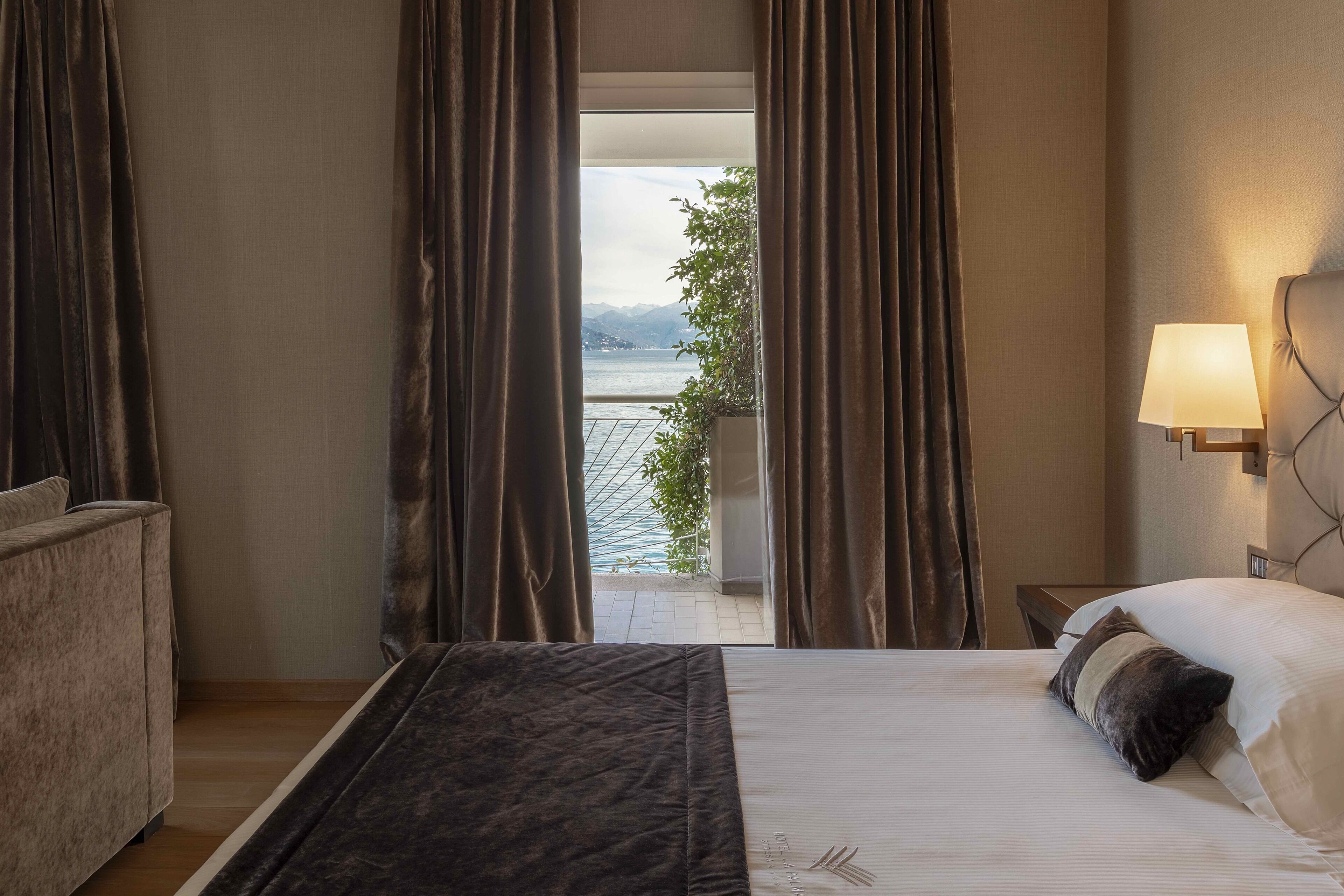 Habitación de hotel con una cama, una mesita de noche con lámpara, y vista a un cuerpo de agua y montañas a través de ventanas amplias.