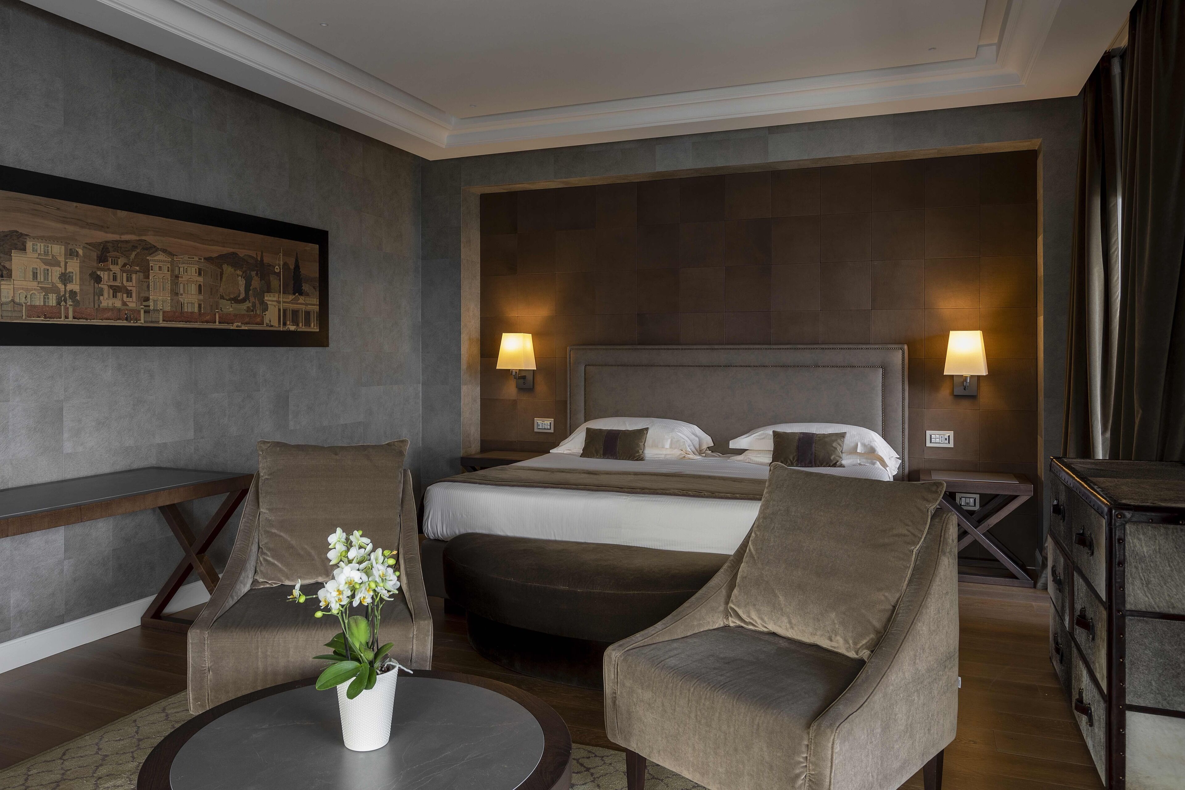 Une chambre d’hôtel avec un grand lit, deux fauteuils, une petite table ronde ornée d’un vase de fleurs et un tableau accroché au mur.