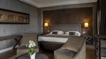 Une chambre d’hôtel avec un grand lit, deux fauteuils, une petite table ronde ornée d’un vase de fleurs et un tableau accroché au mur.