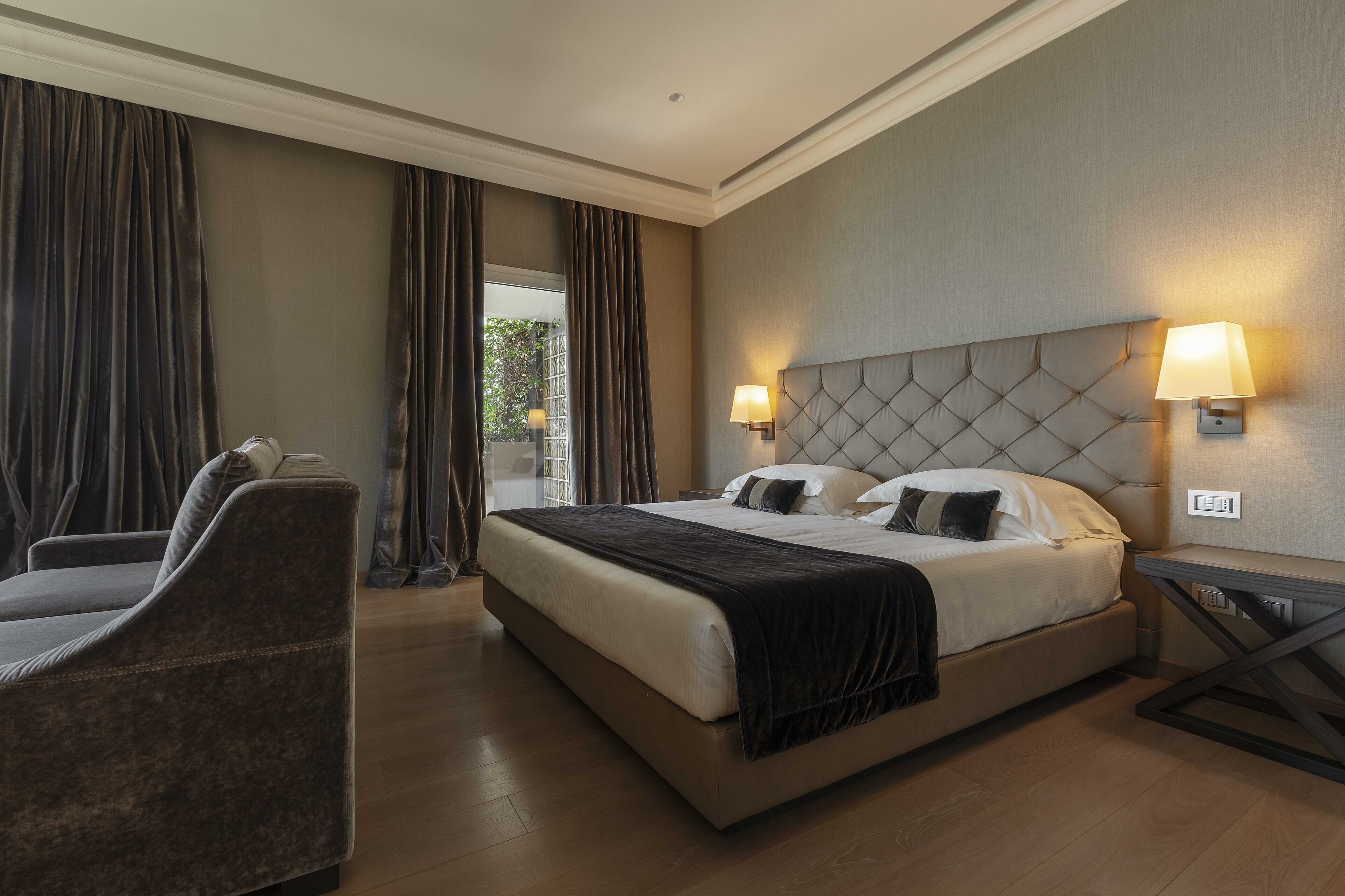 A hotel room with a large bed, two bedside lamps, a chair, and a view of greenery outside.
