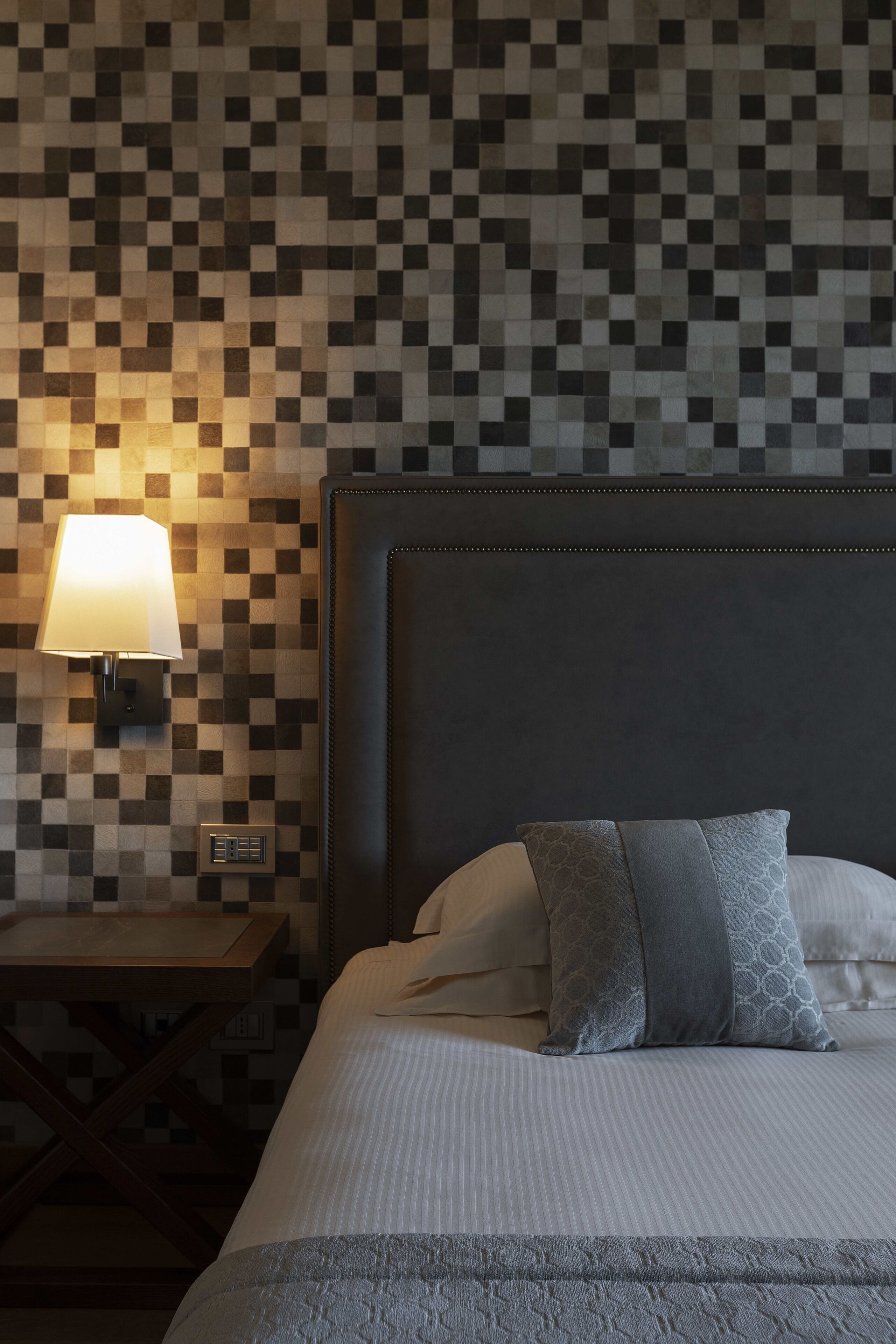 Una cama bien hecha con cabecera gris, una lámpara de pared y una mesita de noche de madera.