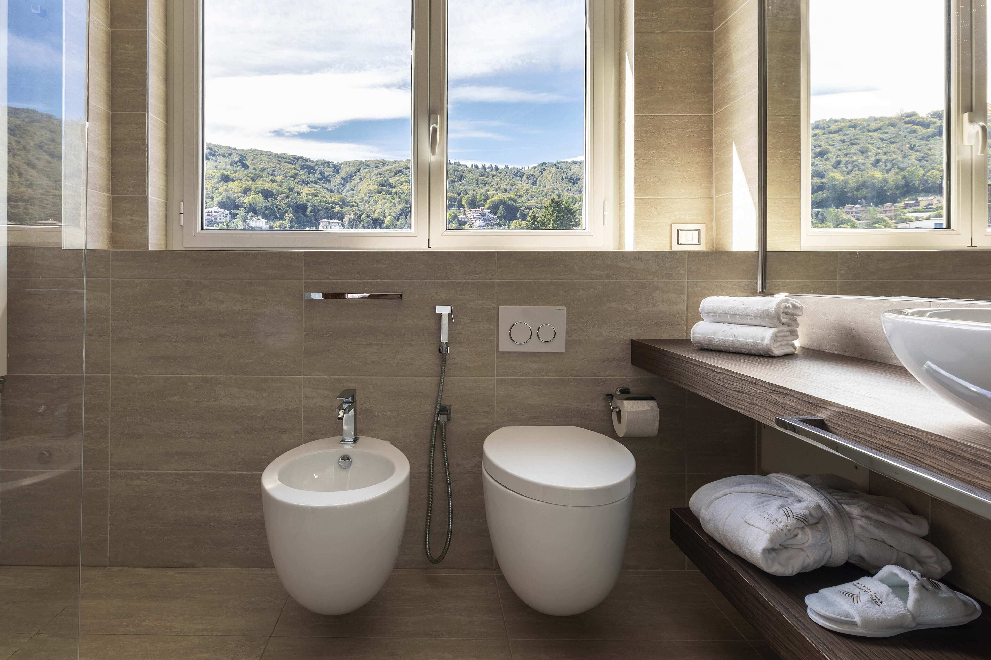 A modern bathroom with a bidet, a toilet, and a large window offering a view of a forested hillside.