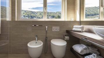 A modern bathroom with a bidet, a toilet, and a large window offering a view of a forested hillside.