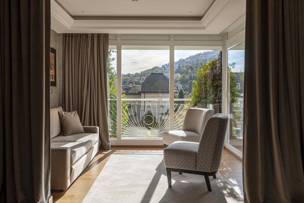 Ein modernes Wohnzimmer mit einer Couch und einem Sessel, großen Schiebeglastüren zu einem Balkon sowie Blick auf Stadt und Hügellandschaft.