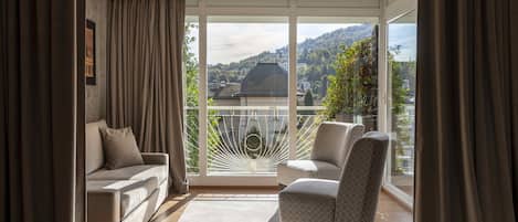 A modern living room with a sofa and armchair, large sliding glass doors to a balcony, and a view of a cityscape and hills.