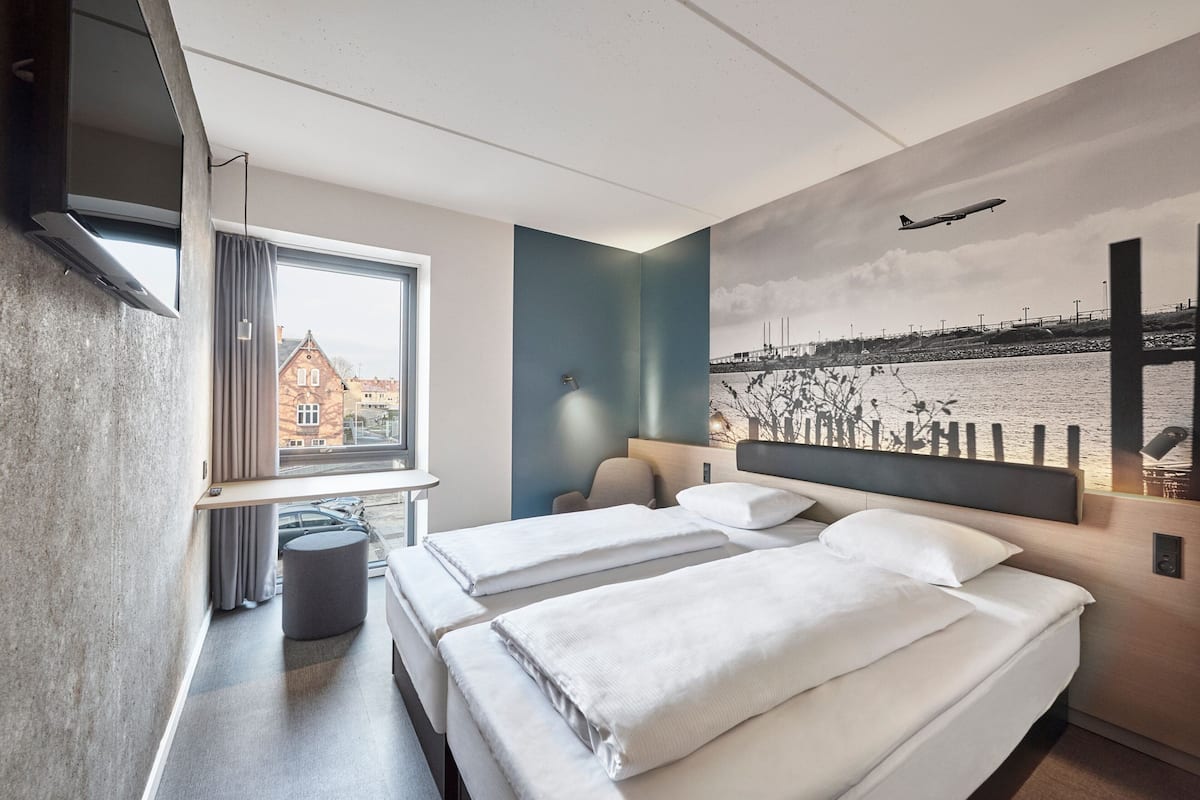 Une chambre d’hôtel avec deux lits, une télévision, une fenêtre avec des rideaux, une lampe et une fresque représentant un pont et une rivière.