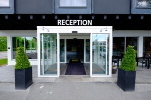 Zona de recepción con puerta de cristal, un cartel encima y dos plantas en macetas a cada lado.