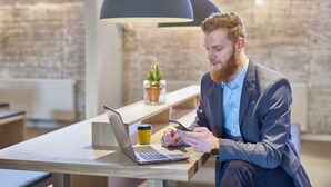 A person sitting at a wooden table with a laptop and a coffee cup, in an office setting with a brick wall and pendant lighting. - Four Points Flex by Sheraton Copenhagen Airport (Kastrup)