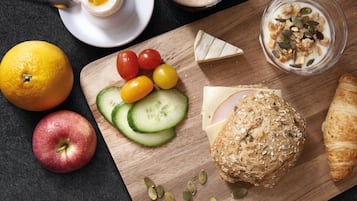 Een houten plank met plakjes komkommer, cherrytomaatjes, een appel, een plak kaas, een broodje en een glas yoghurt met zaden.