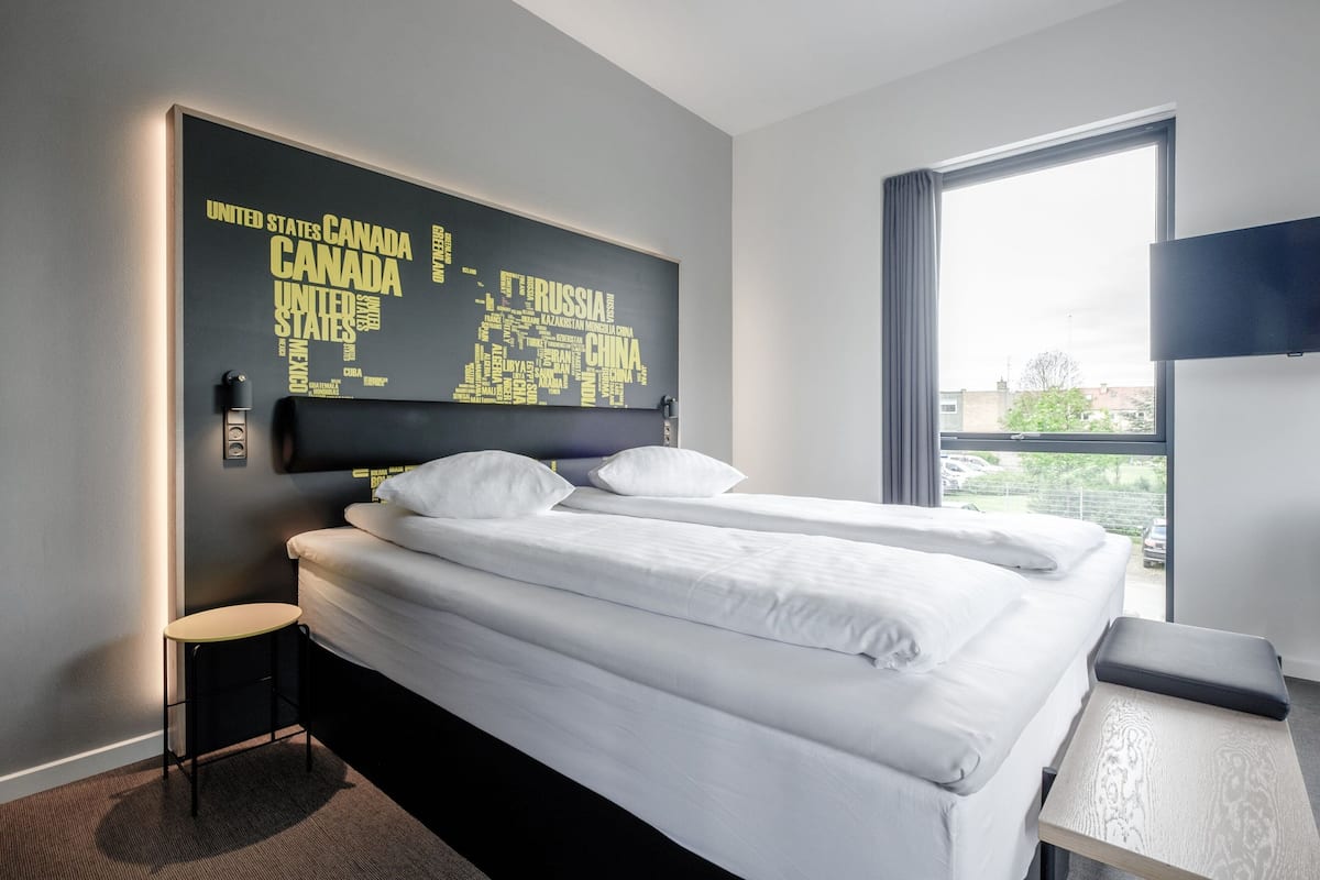 Une chambre d’hôtel avec un lit, un petit tabouret, une table et une grande fenêtre.