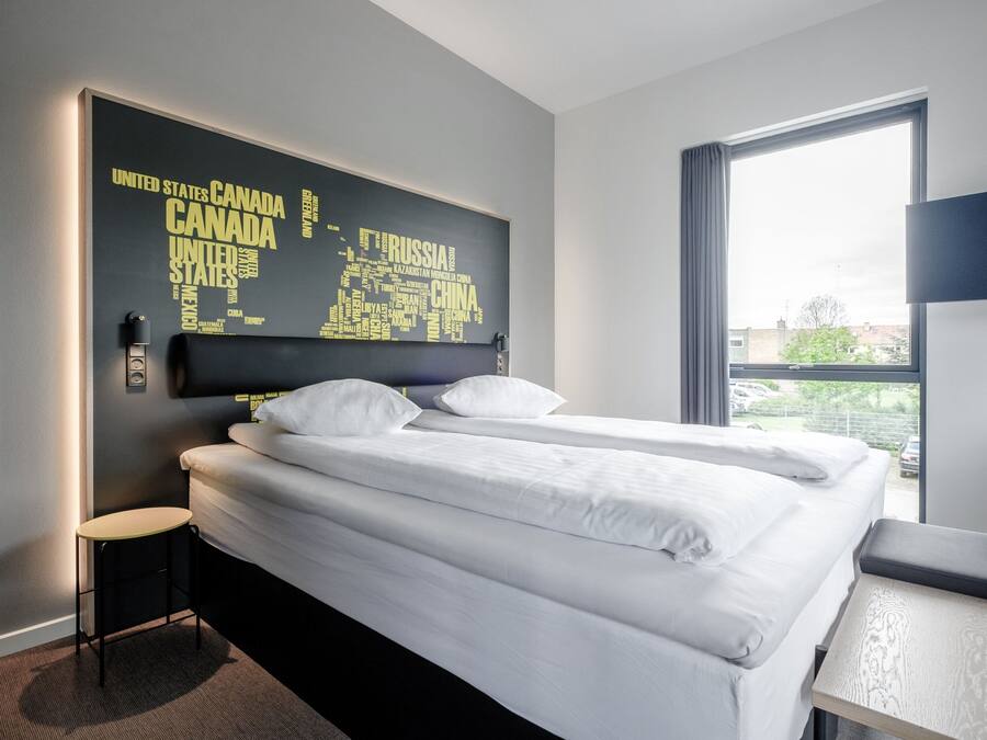 Ein Hotelzimmer mit Bett, einem kleinen Hocker, einem Tisch und einem großen Fenster.
