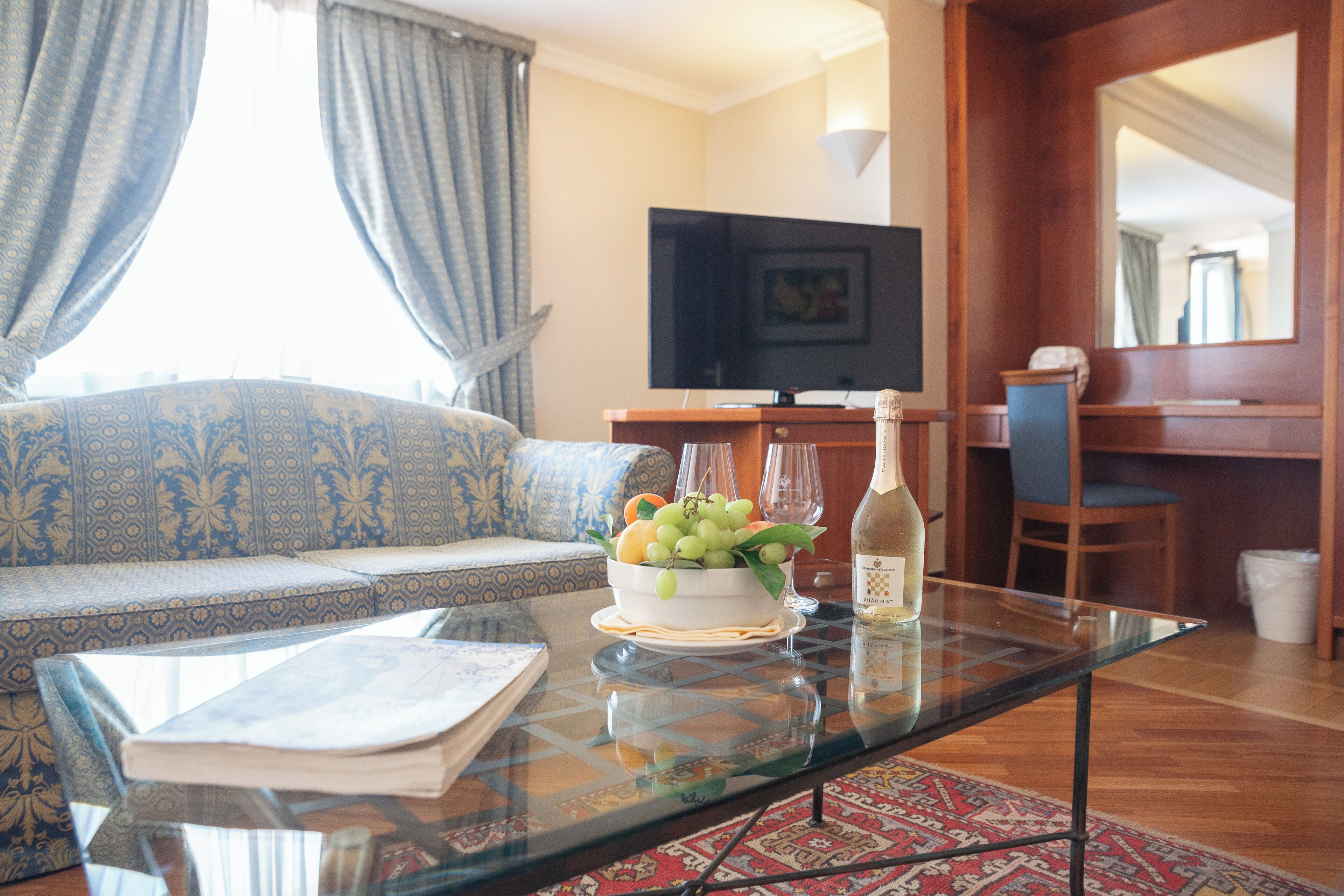A living room with a sofa, a glass coffee table with a fruit bowl and a bottle, a television, and a wooden dining area.