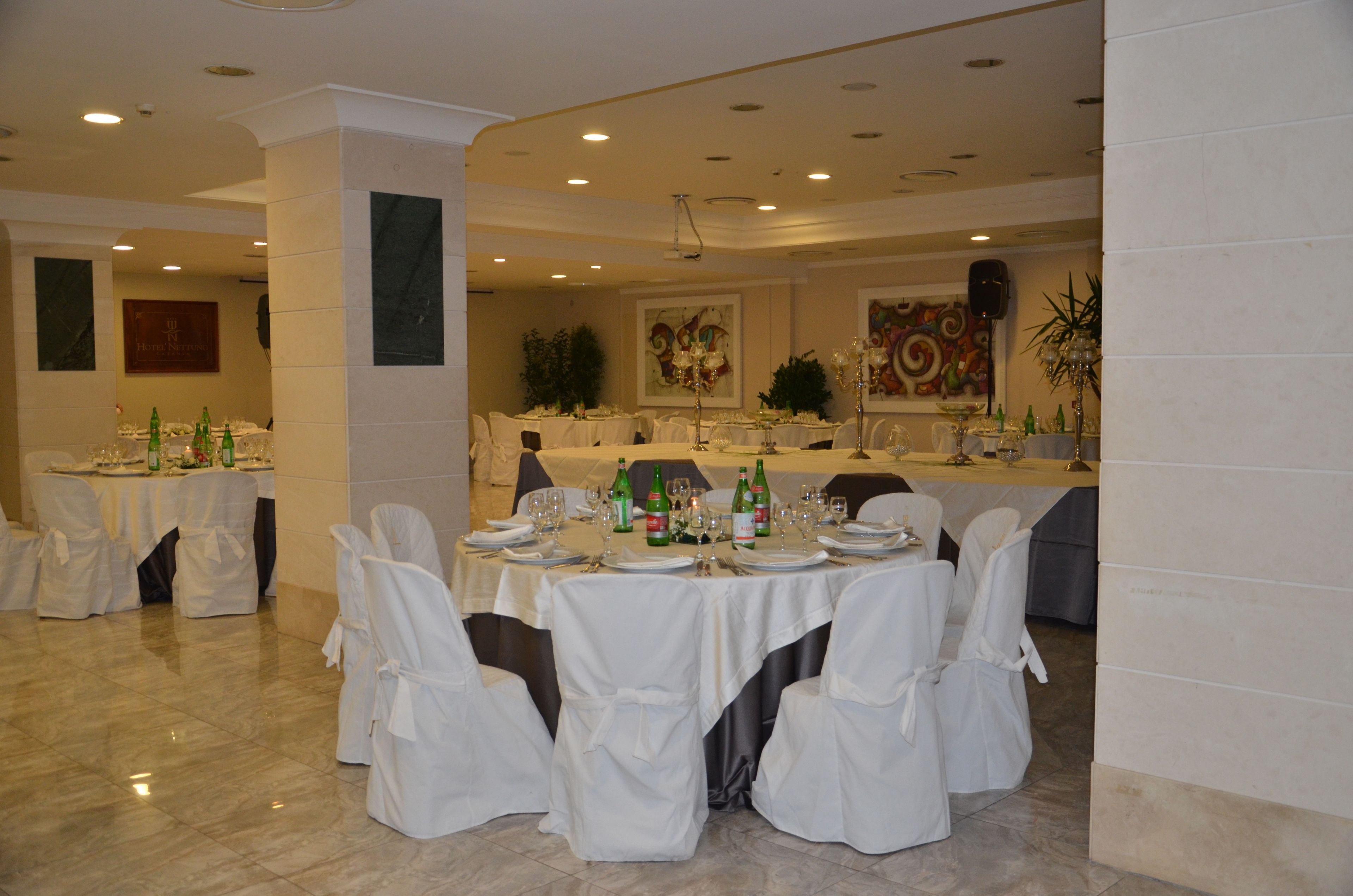 A banquet hall with round tables set for a formal event, featuring white tablecloths and chairs, and a stage with a speaker and decorative plants.