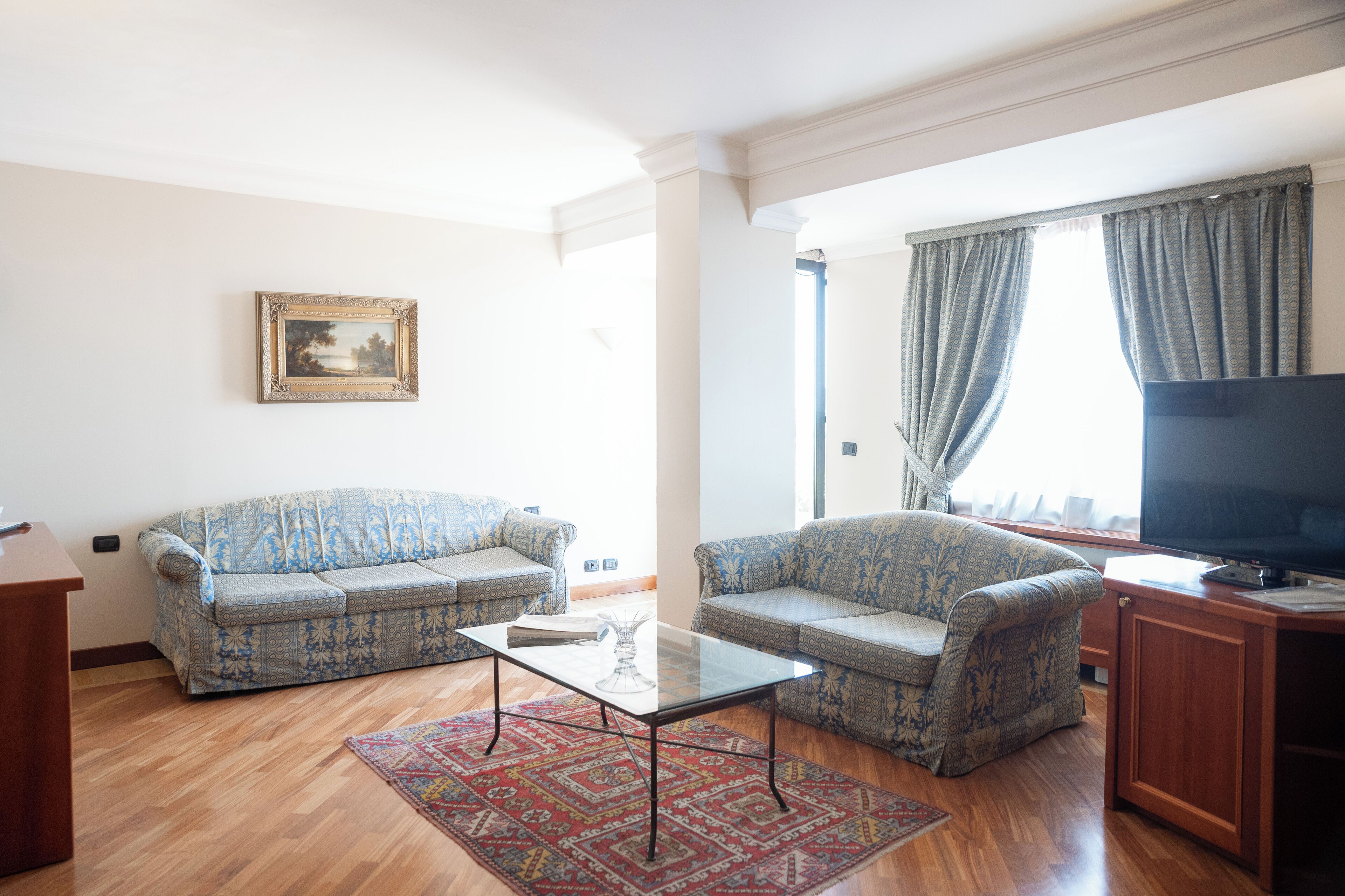 Un salon avec deux canapés à motifs, une table basse en verre, un meuble en bois, un téléviseur à écran plat et un tableau encadré accroché au mur.