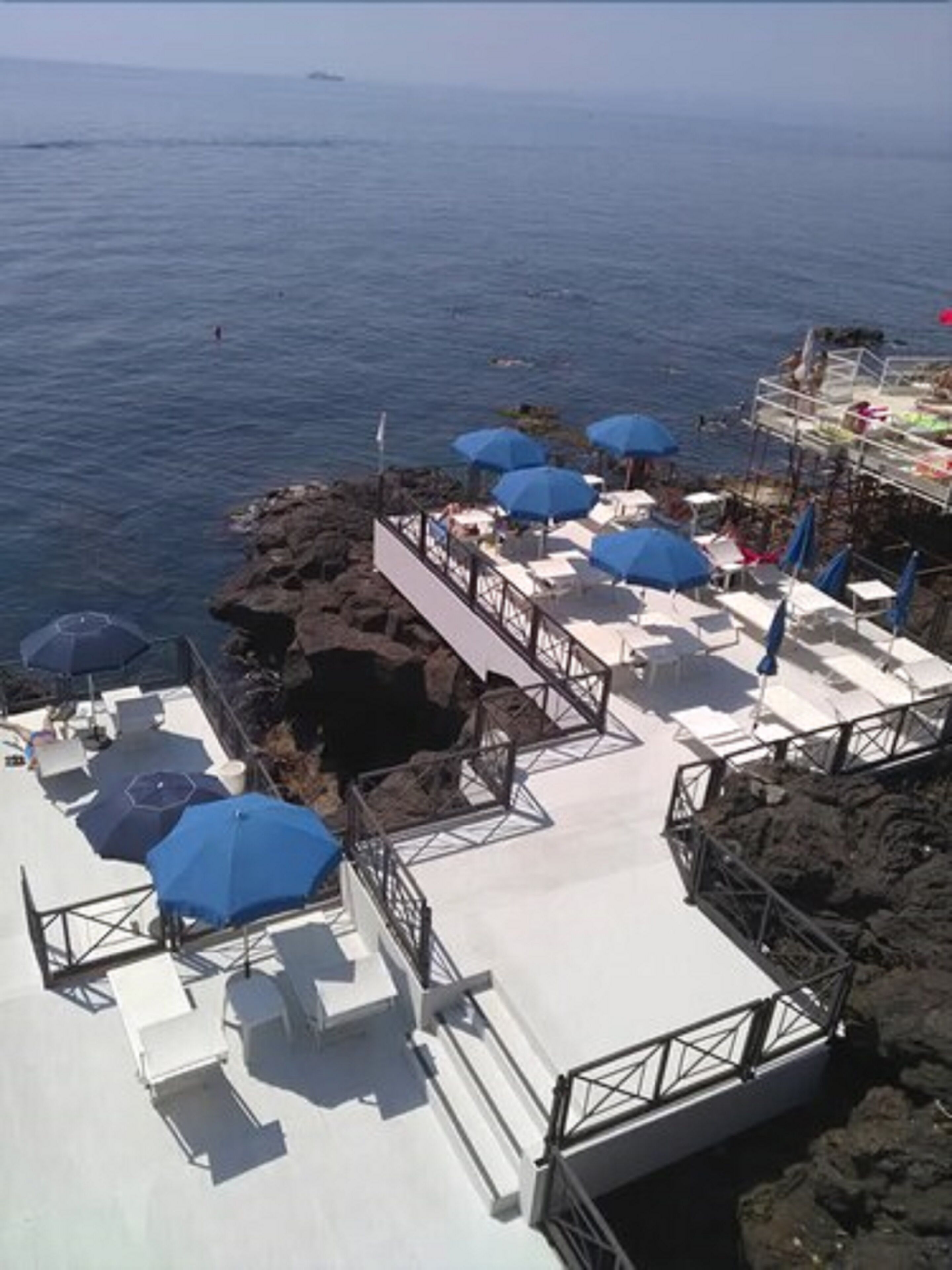 A seaside terrace with white lounge chairs and blue umbrellas, overlooking the ocean.