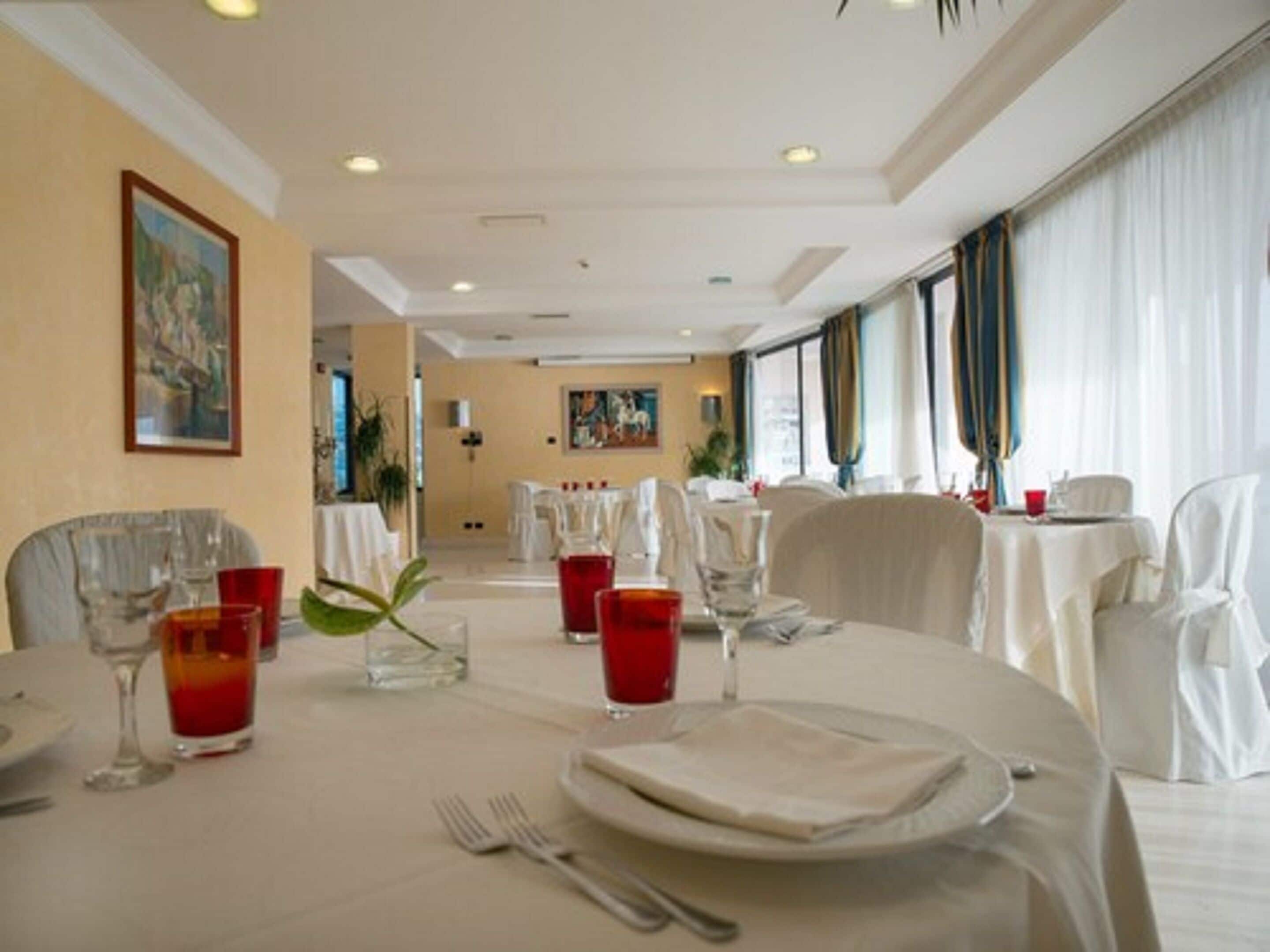 A dining area with round tables set for four, each with white tablecloths, red glasses, and silverware. The room has cream-colored walls, a large window with curtains, and a painting on the wall.