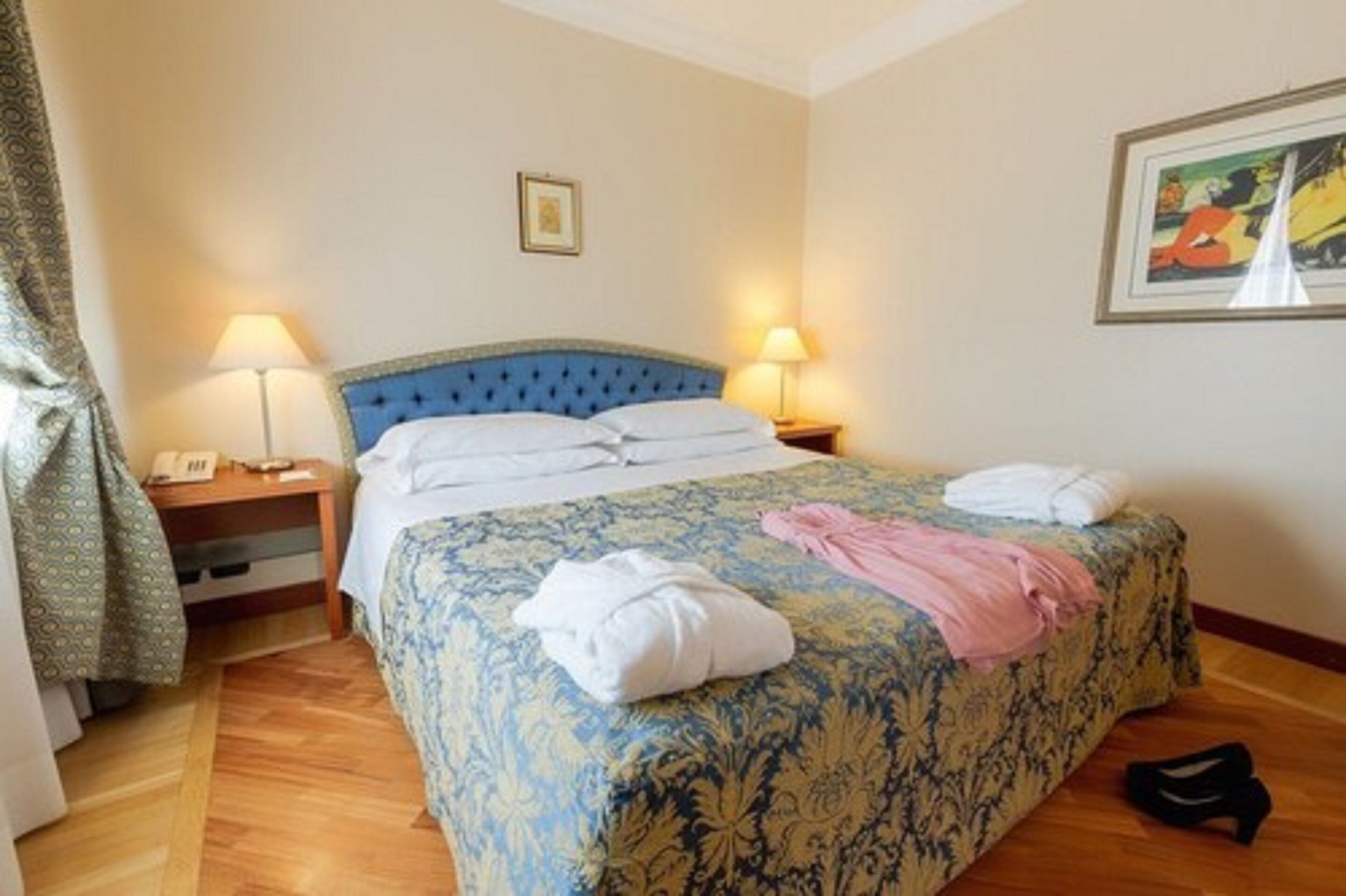 A hotel room with a large bed, two bedside lamps, a framed picture on the wall, and a pair of shoes on the floor.