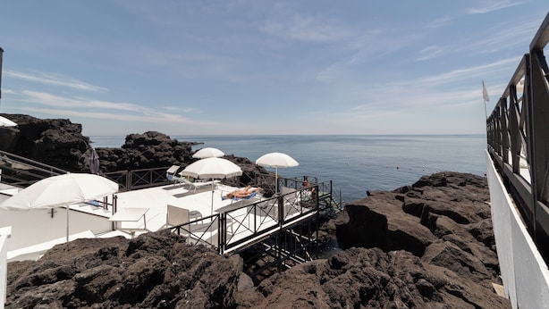 A rocky coastal area with white umbrellas and lounge chairs on a deck overlooking the sea. - Hotel Nettuno (Catania)