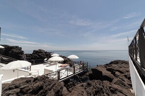 A rocky coastal area with white umbrellas and lounge chairs on a deck overlooking the sea. - Hotel Nettuno (Catania)