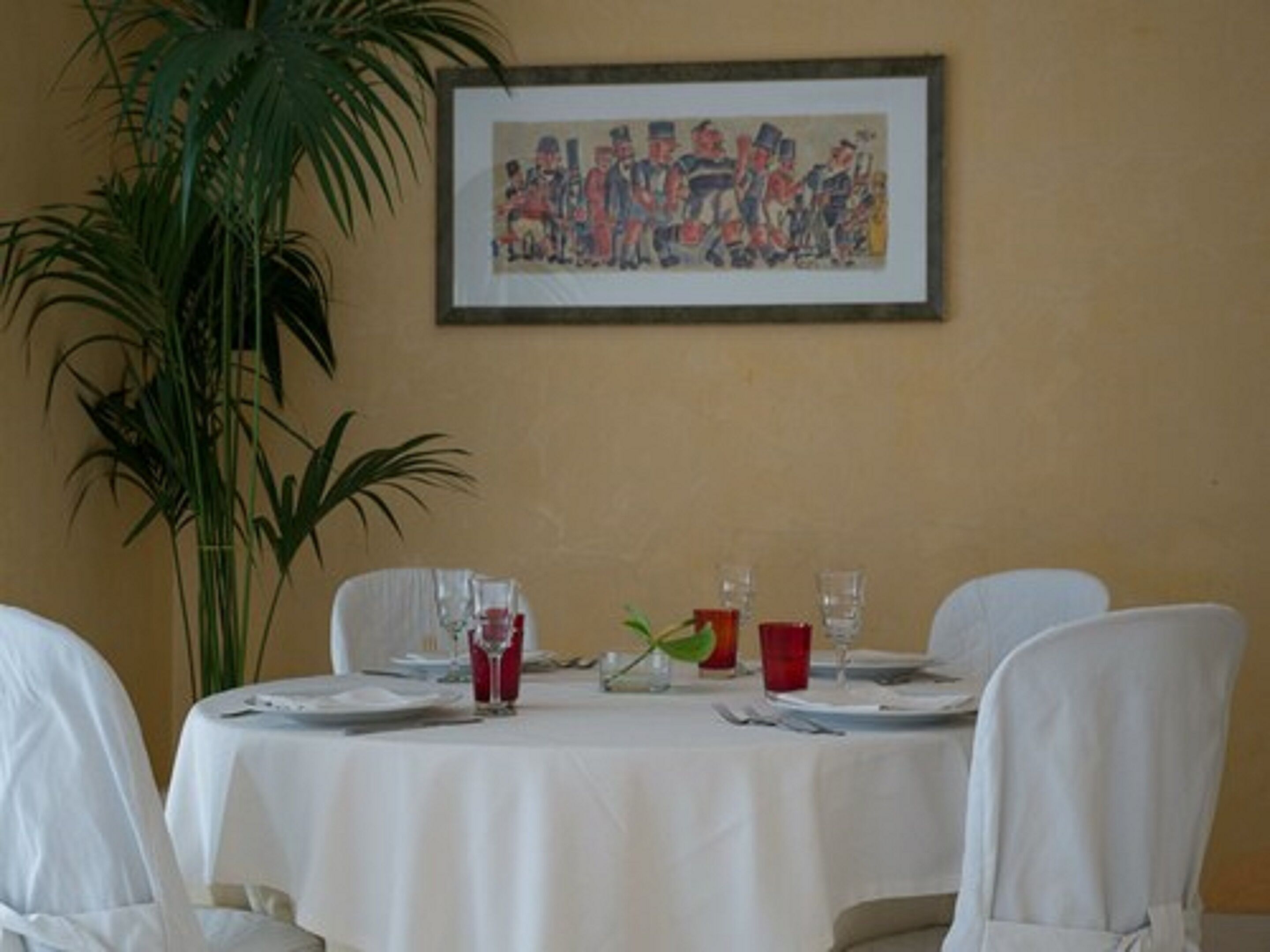 A dining table set for four with white tablecloths, red and clear glasses, and a framed painting on the wall.