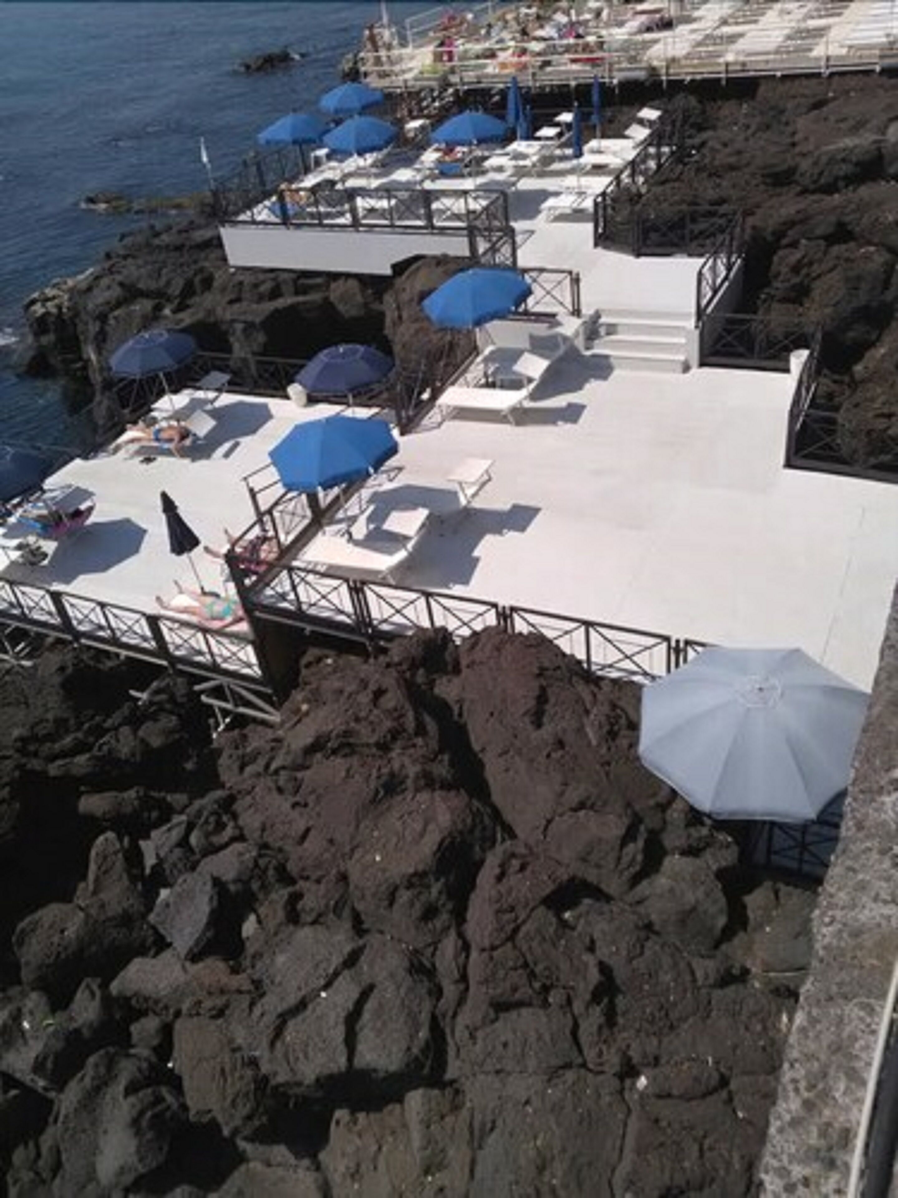 Zona costera rocosa con sillas de playa y sombrillas, una escalera que baja al agua y una barandilla con vistas al mar.