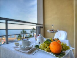 Una mesa con mantel blanco, una taza y platillo, un plato con uvas y una naranja, y un pequeño soporte para postres con pasteles.
