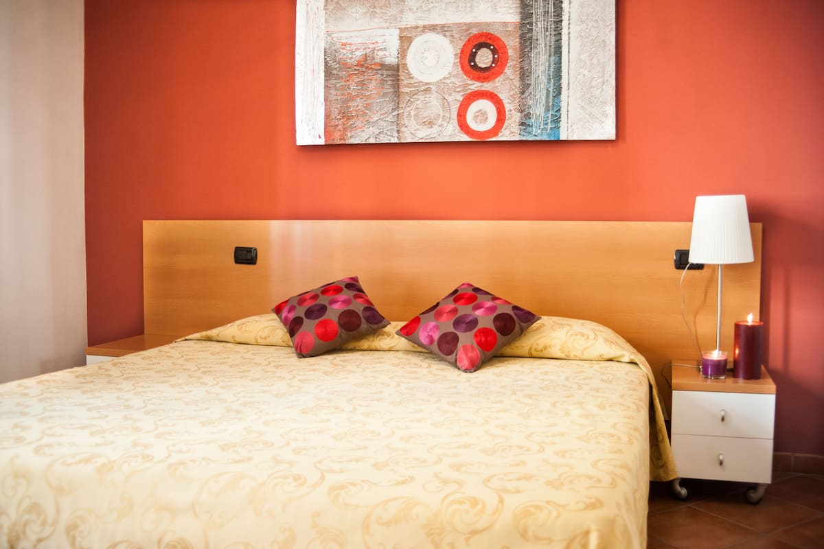 A neatly made bed with patterned bedding, a bedside table with a lamp and candles, and a wall art piece above the bed.