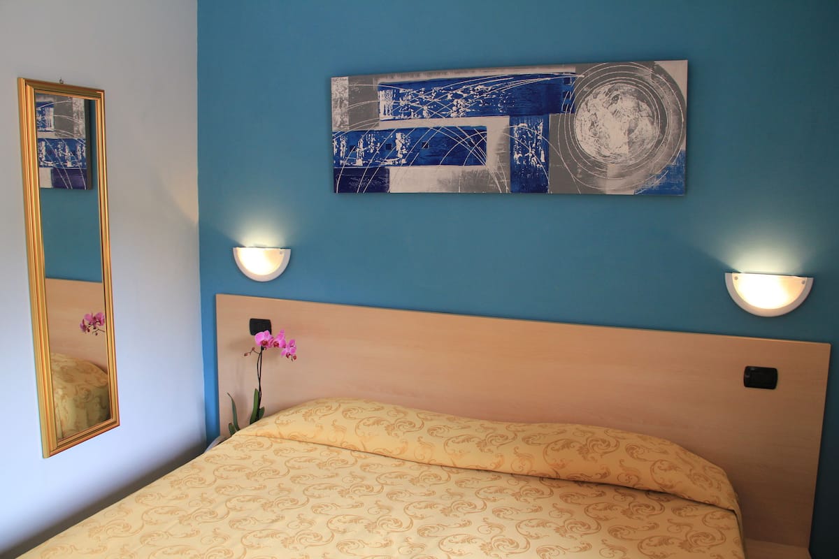 A bedroom with a bed, a wall-mounted painting, two wall sconces, and a mirror.