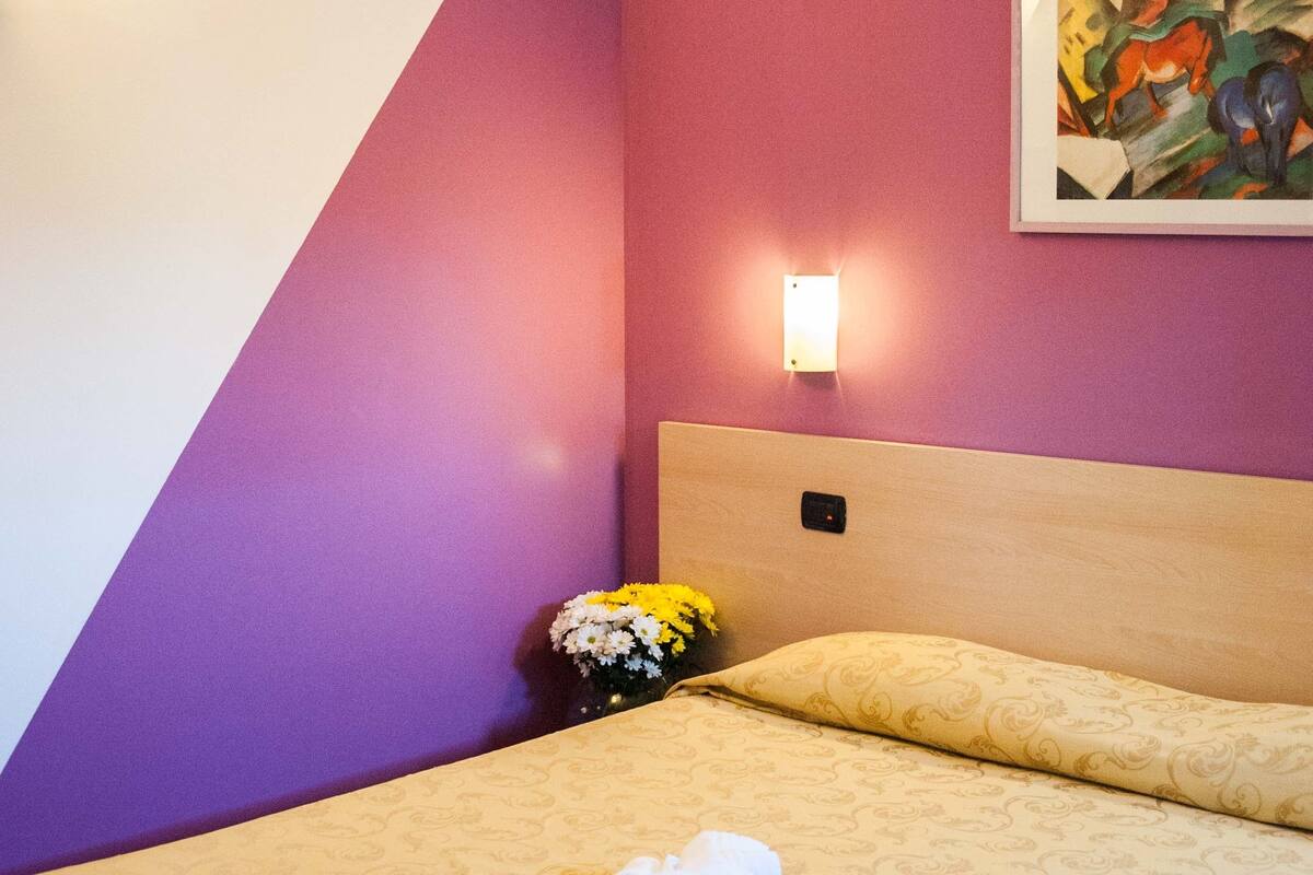 A hotel room with a bed, a nightstand, a wall-mounted light, and a painting.