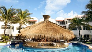 A pool with a thatched-roof bar area, surrounded by palm trees and a building with white walls and red roofs.