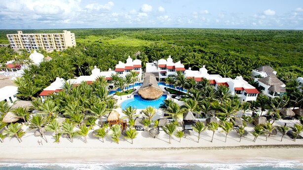 A resort with a central pool, surrounded by palm trees and beach huts, overlooking a beach and forested area.